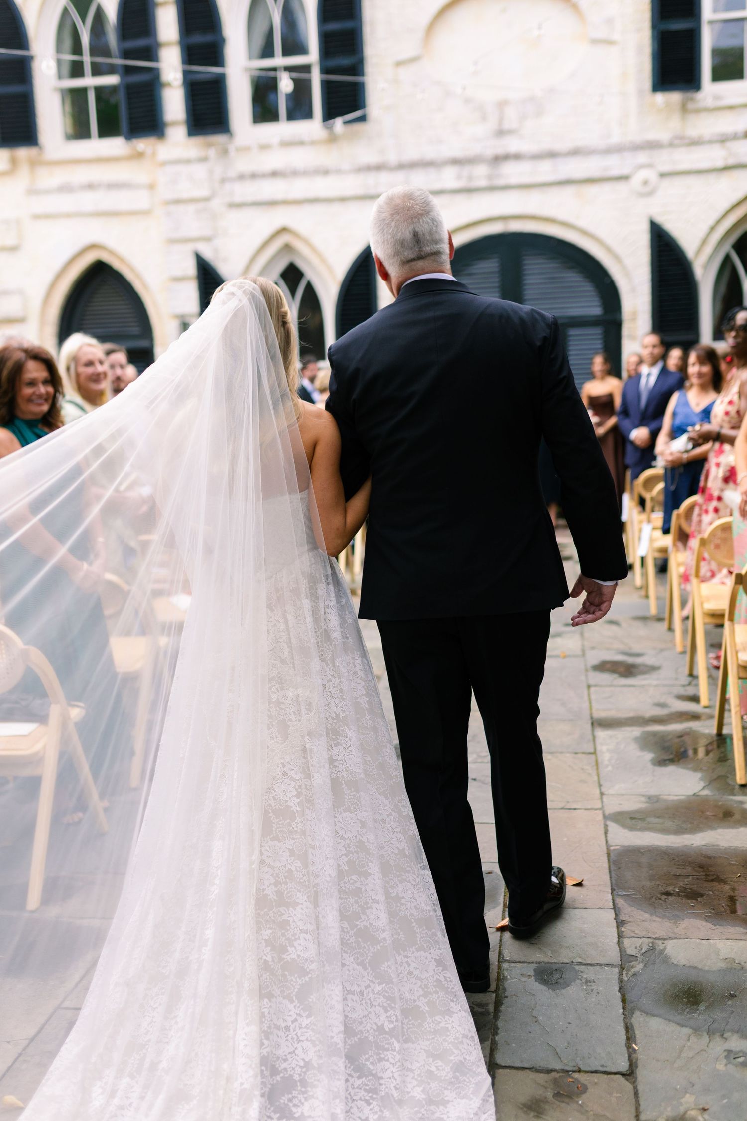 Bride and father walking down the aisle at an outdoor wedding ceremony, bride wearing a long lace veil.