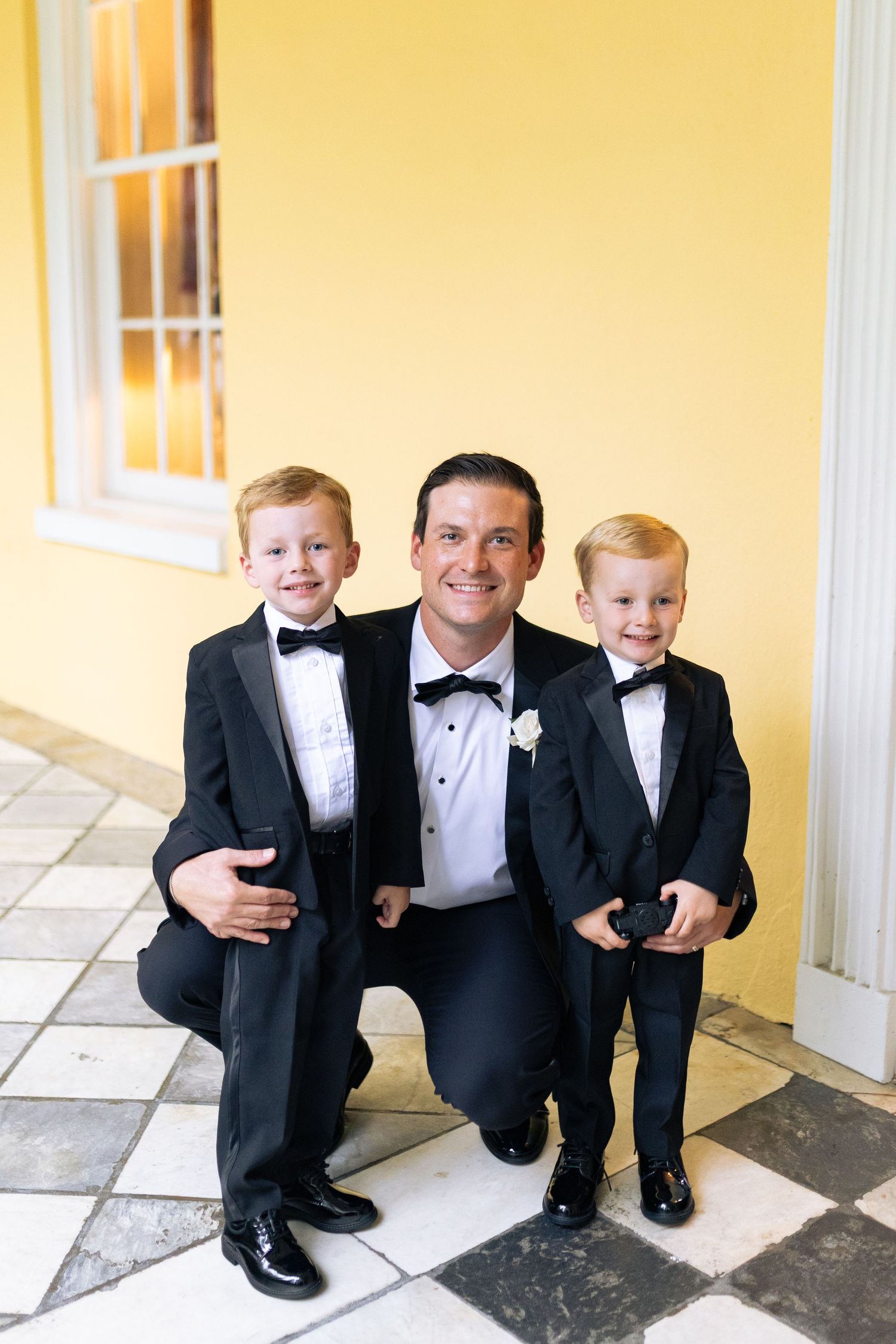 Young ring bearers in tuxedos sit together on a tiled floor.