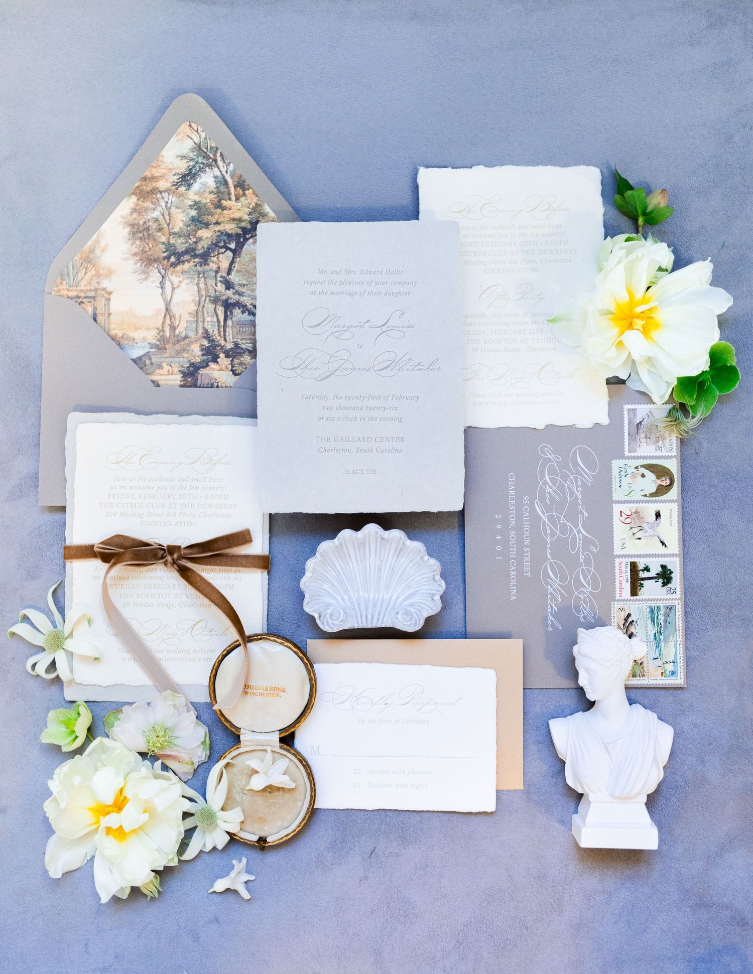 Elegant wedding invitation suite with gray envelopes, floral liner, white flowers, shell dish, and ring box on a blue backdrop.