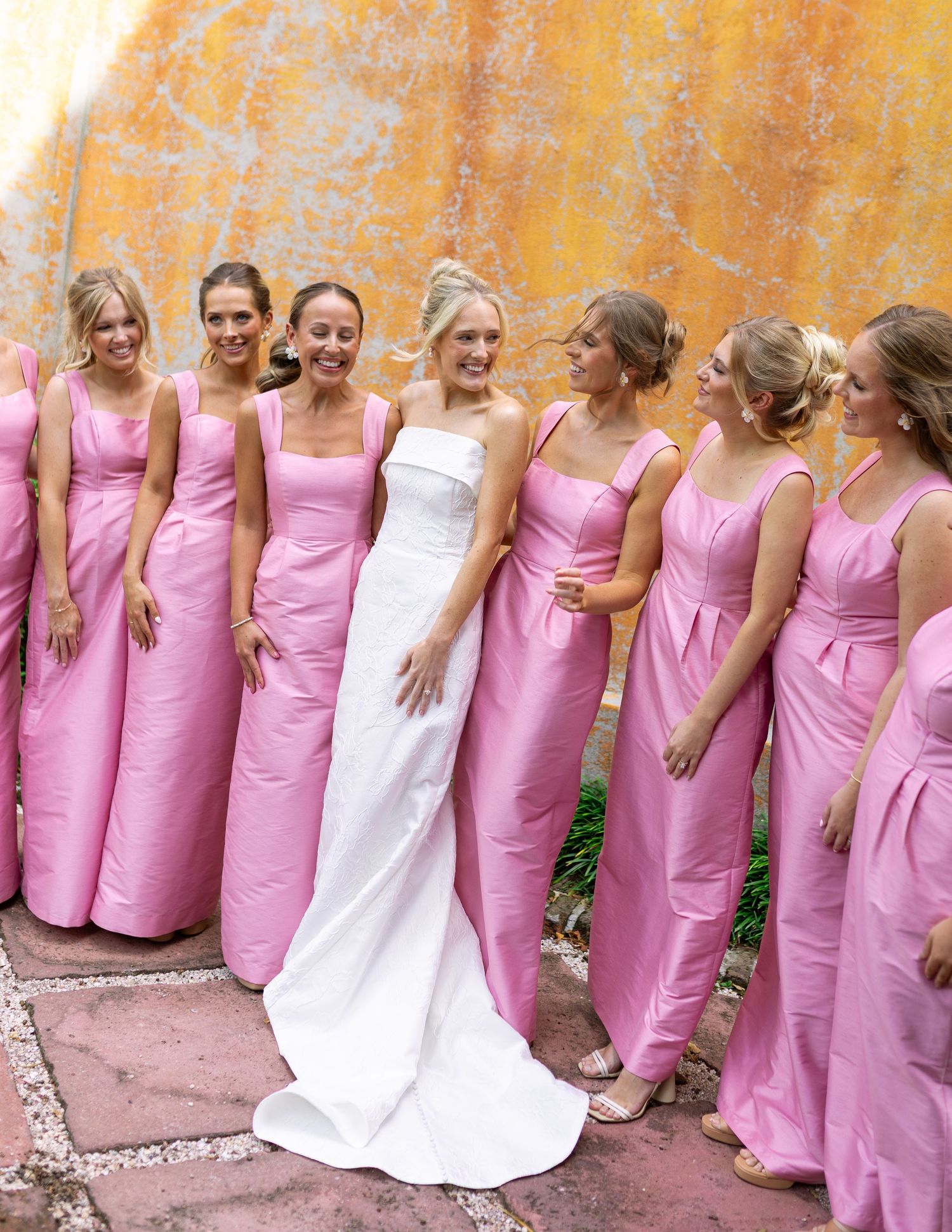 Wedding party lined up against a yellow wall wearing pink dresses with bride in white gown.