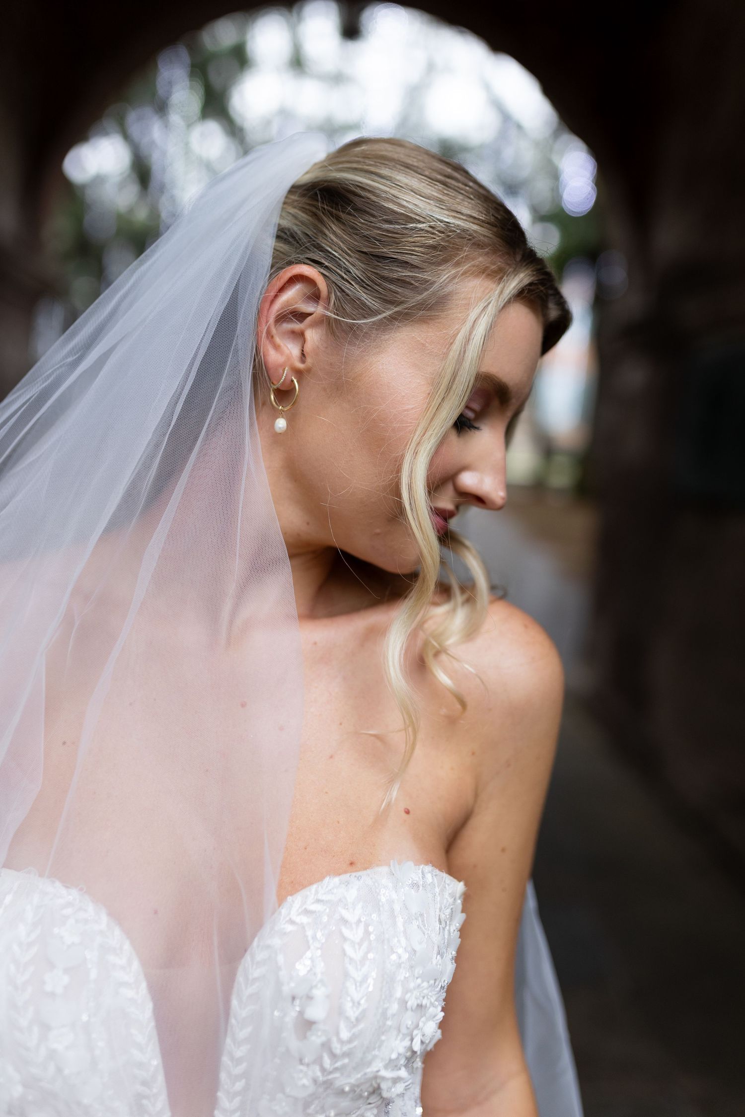 Side profile view of a bride wearing a white lace gown and veil with pearl earrings.