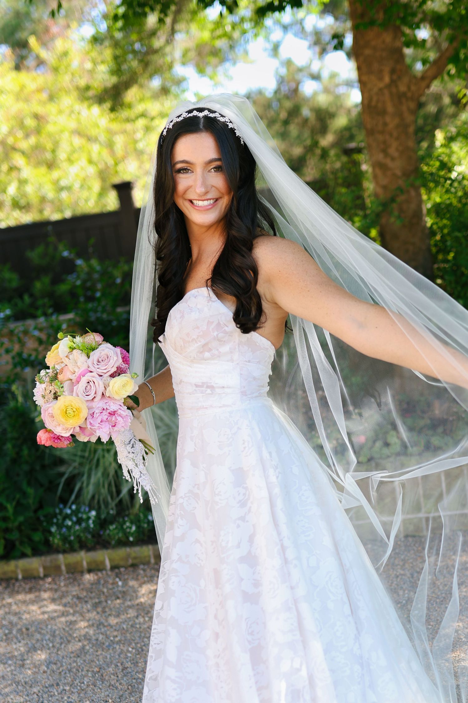 Smiling bride in white lace gown and veil holds colorful bouquet outdoors among green trees on her wedding day.