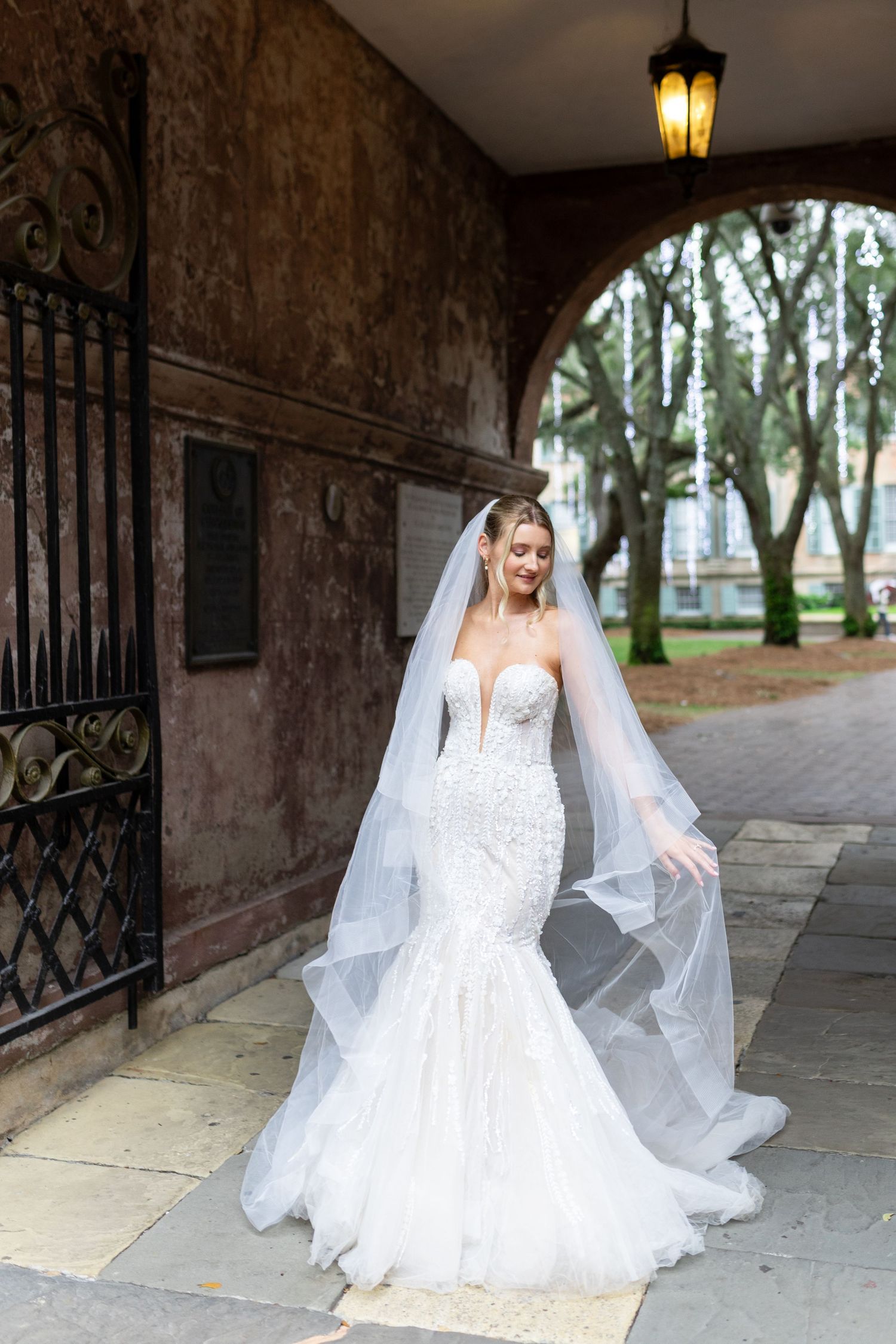 Bride in lace gown poses under historic archway with flowing veil.