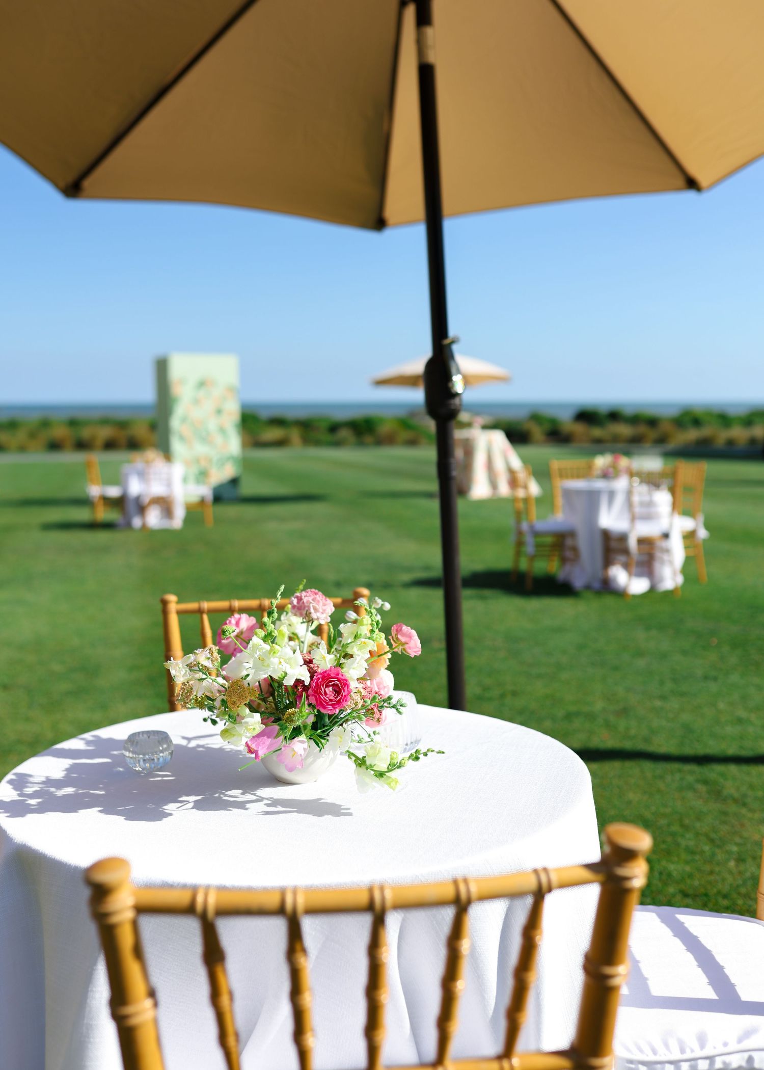 Elegant outdoor wedding reception setup with white tablecloths, floral centerpieces, gold chairs, and a patio umbrella on a lush green lawn.