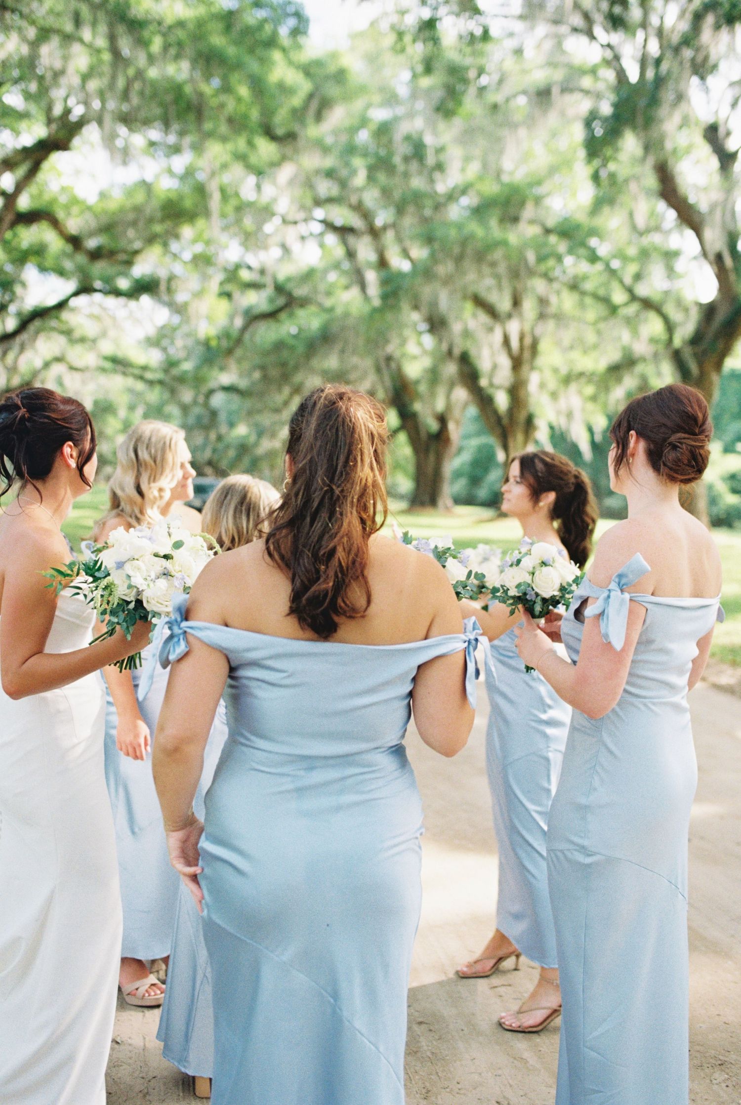 Bridesmaids in light blue off-shoulder gowns with bow details hold white floral bouquets outdoors under lush green trees.
