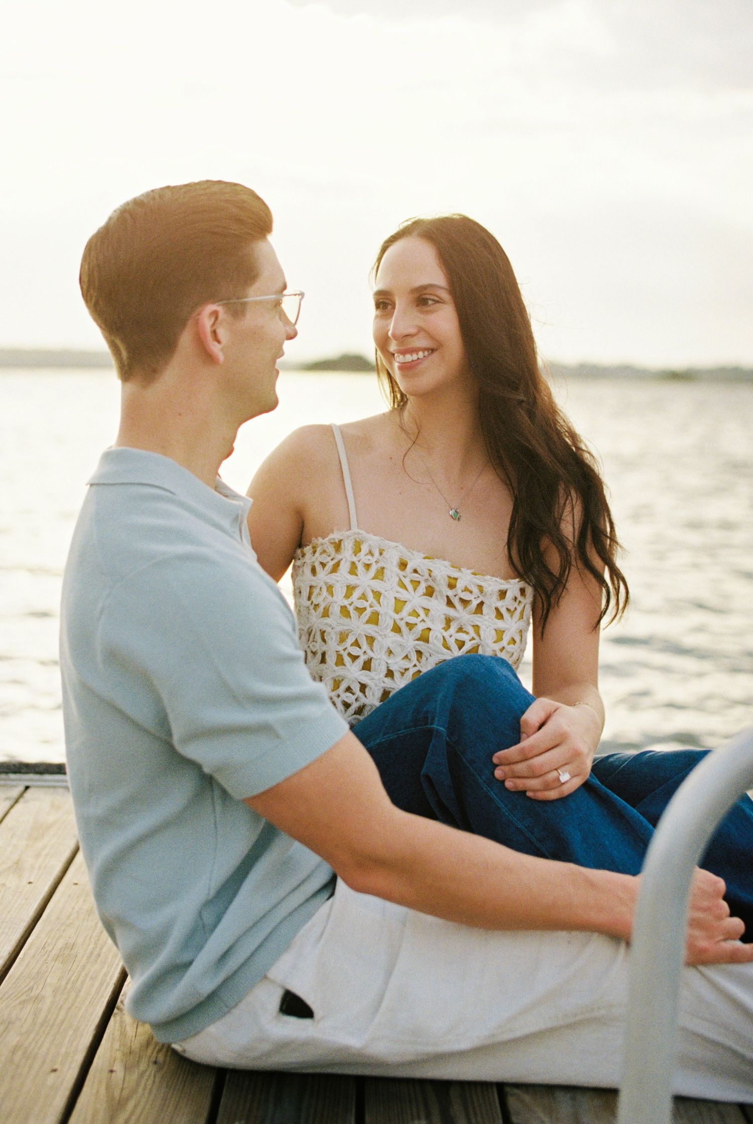 Couple sitting on a wooden dock by the water at sunset, smiling at each other in casual summer outfits.