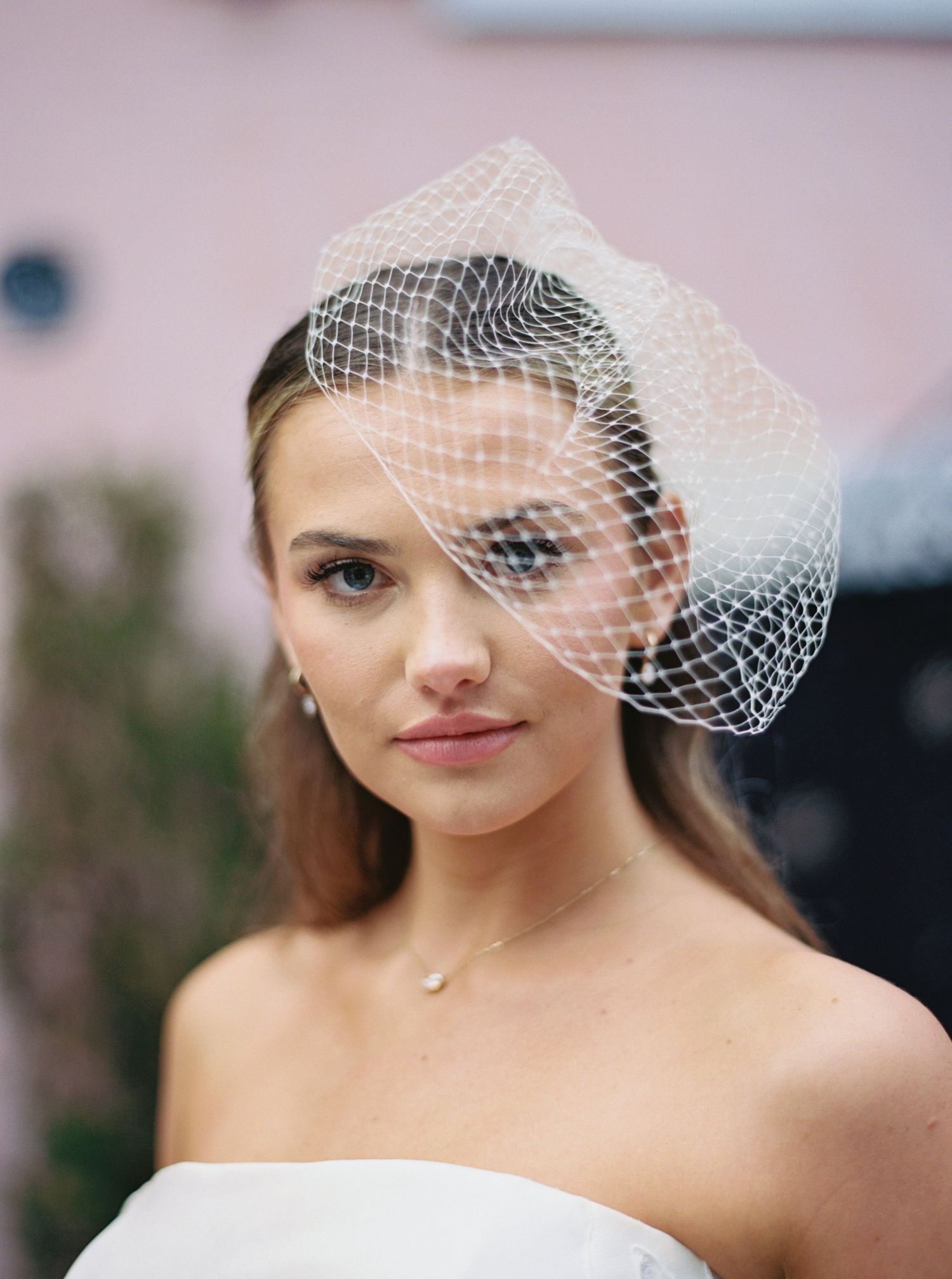 Bride wearing a white birdcage veil with elegant makeup and a strapless gown, posing outdoors.