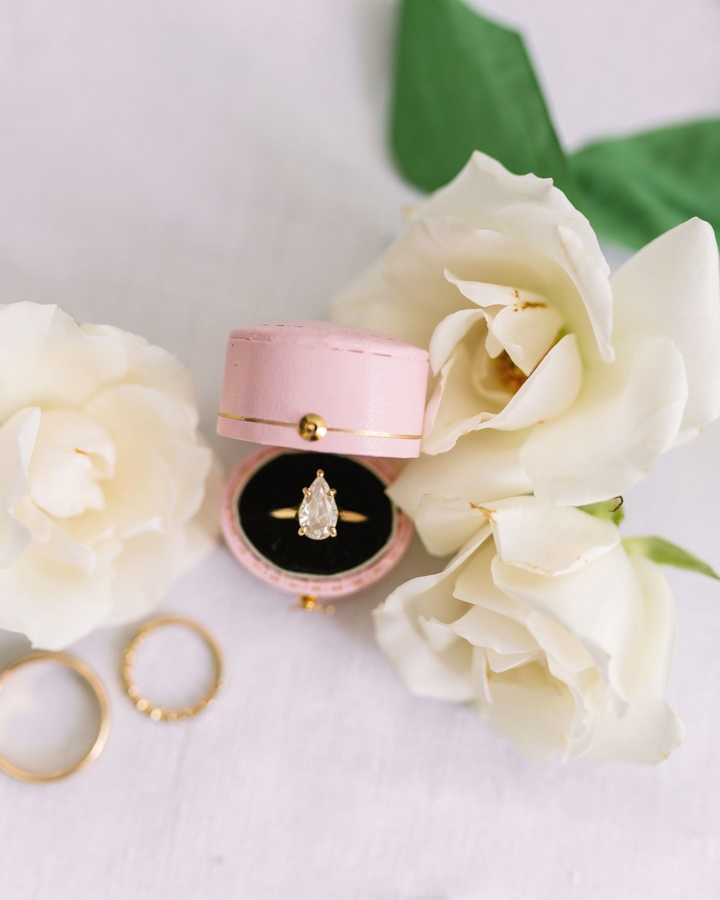 Wedding rings and jewelry displayed with white roses and pink ring box on light background.