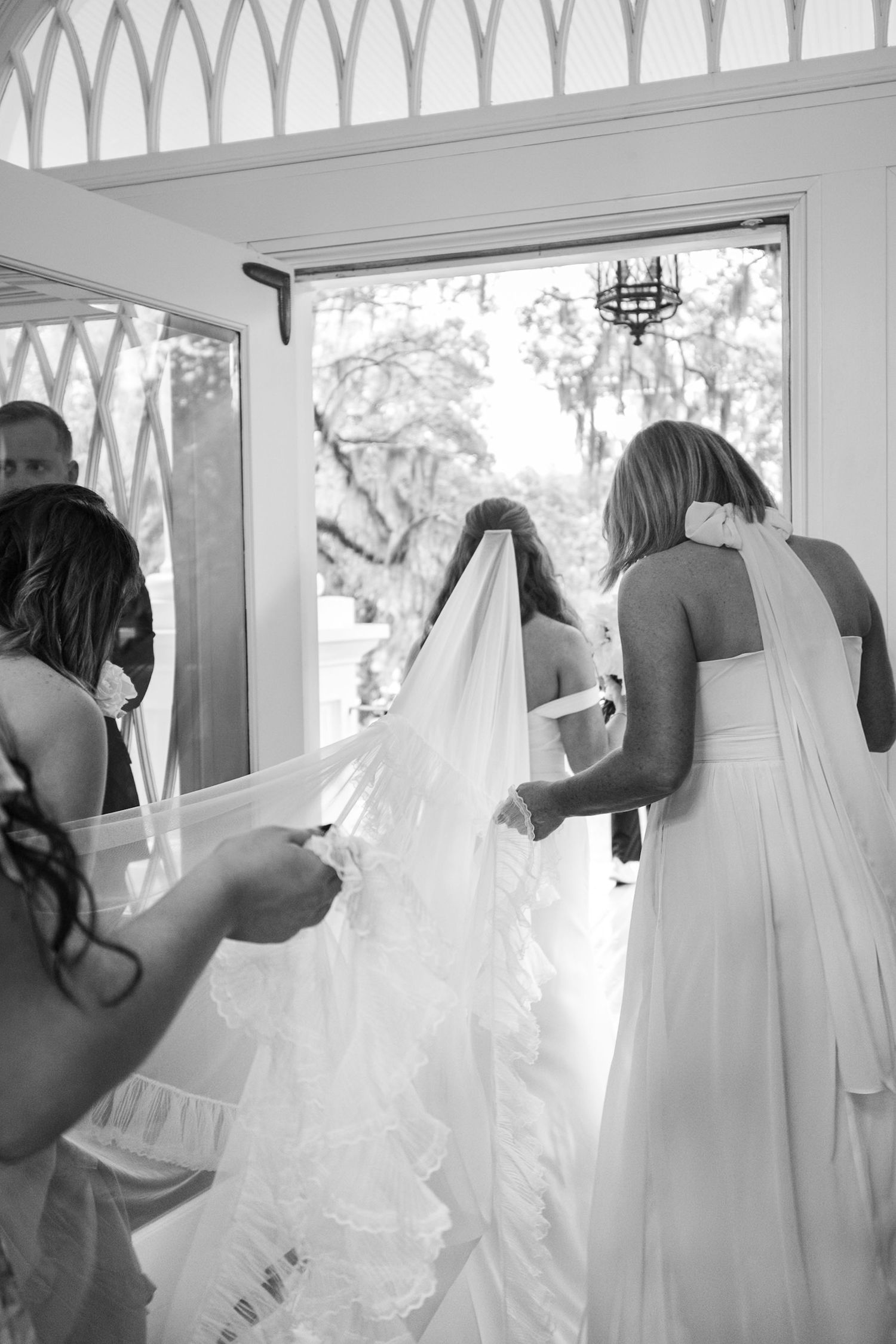 Wedding party helps adjust the bride's dress train through white lattice doorway.
