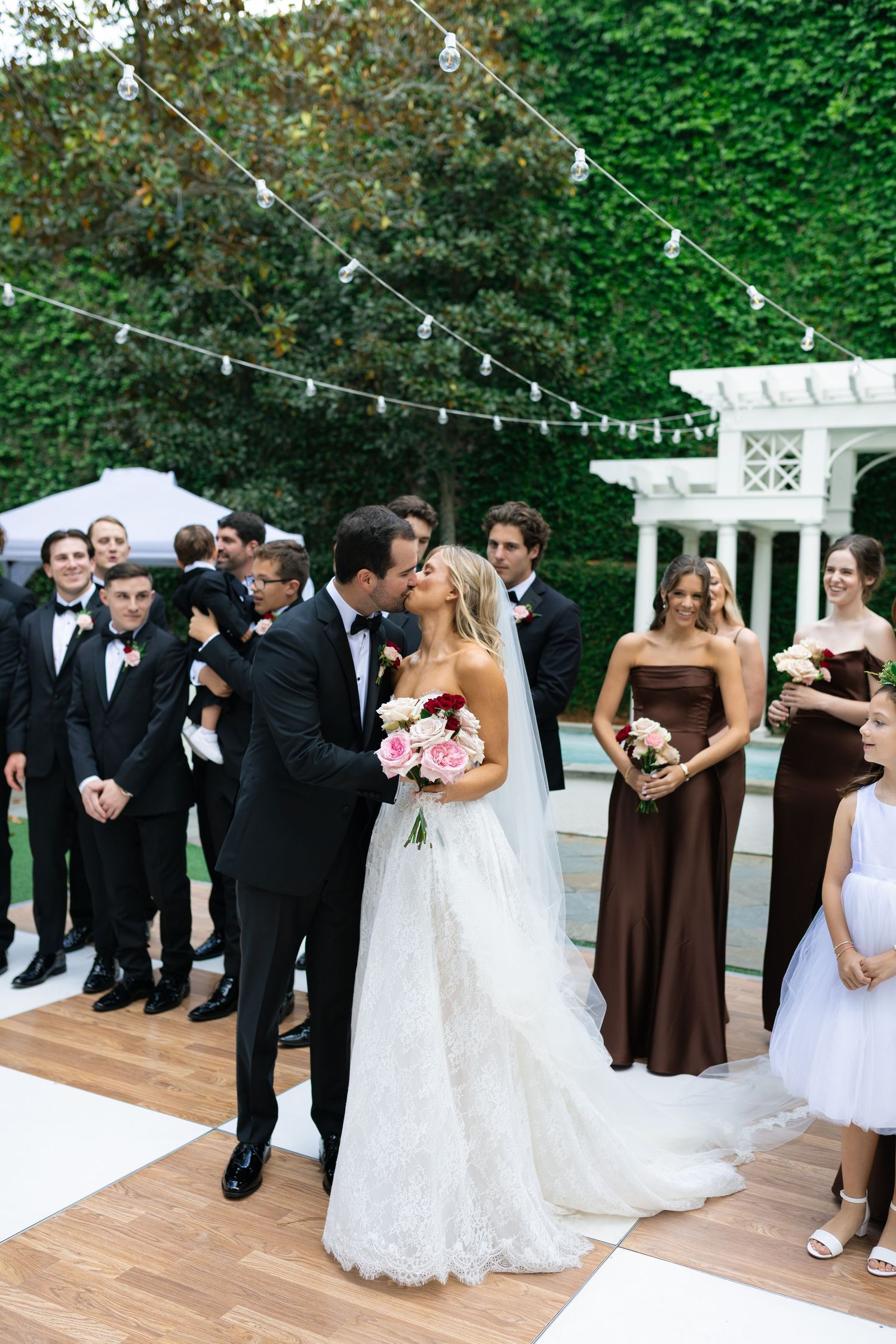 Bride and groom share first kiss at outdoor wedding ceremony with bridal party in black tuxedos and brown bridesmaid dresses.