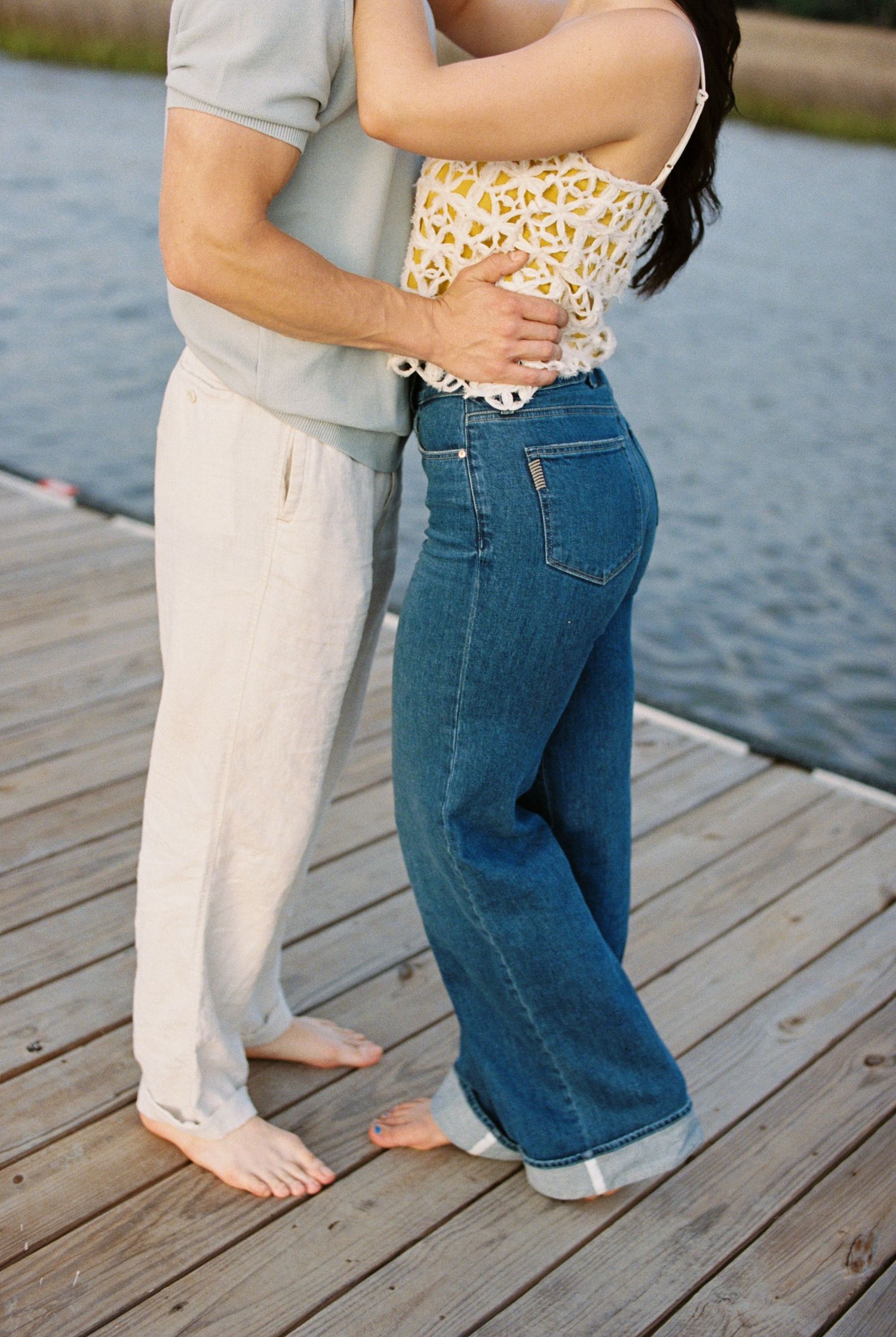 Couple embracing on a wooden dock, wearing linen pants and wide-leg jeans, with water in the background.