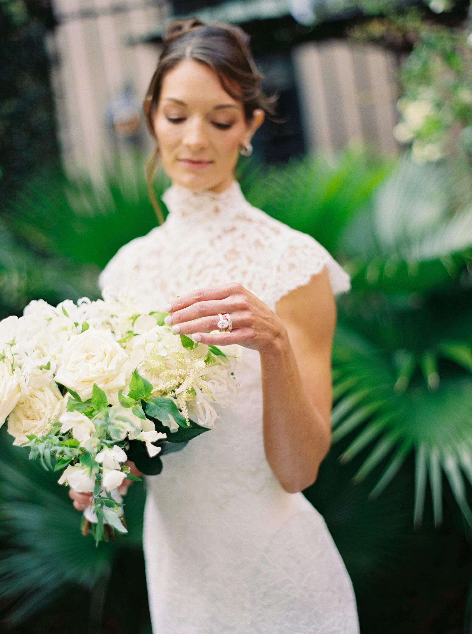 Bride in white lace high-neck gown admires her engagement ring while holding elegant white floral bouquet outdoors.