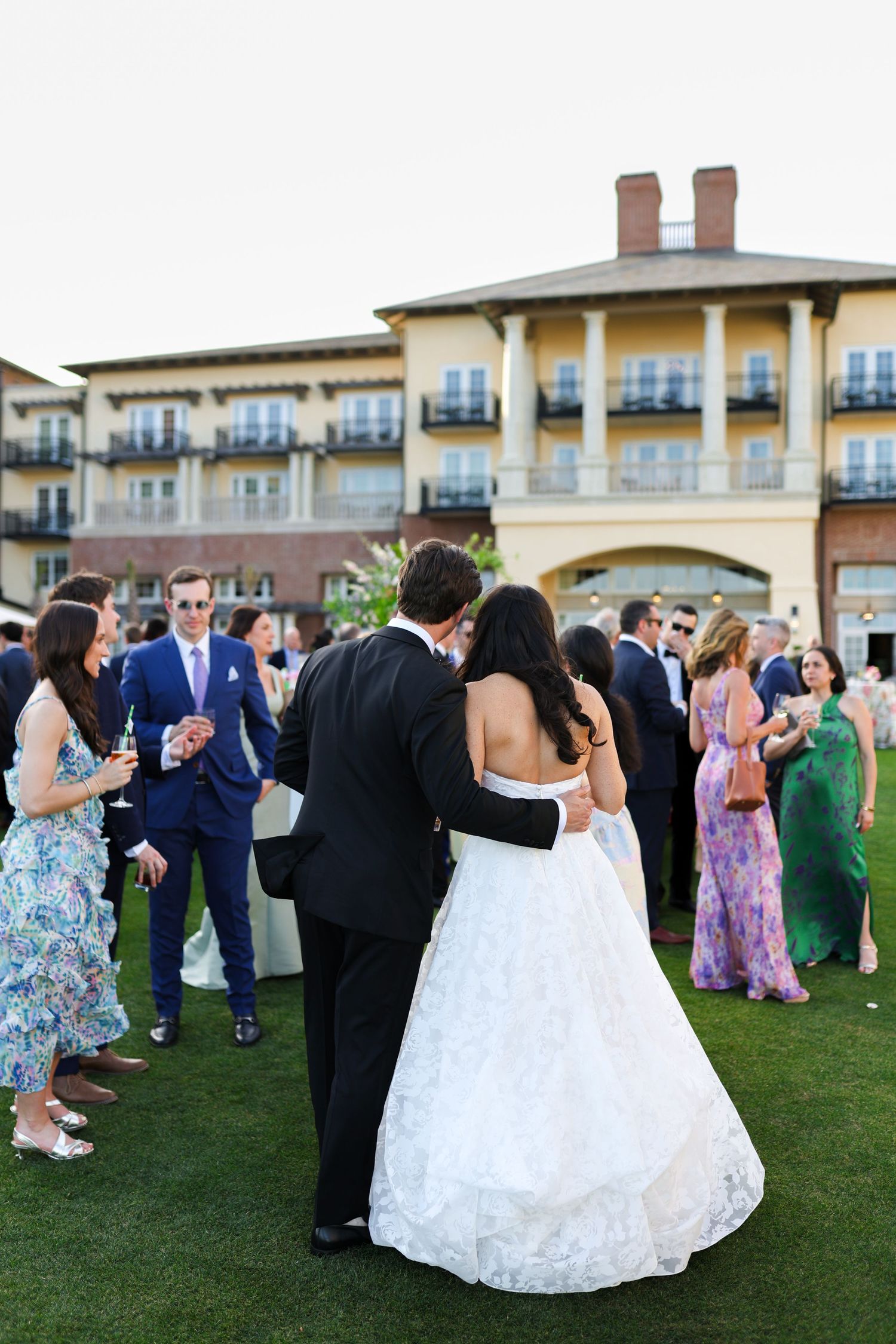 Bride and groom share a romantic moment outdoors at their wedding reception with guests mingling near a grand hotel building.
