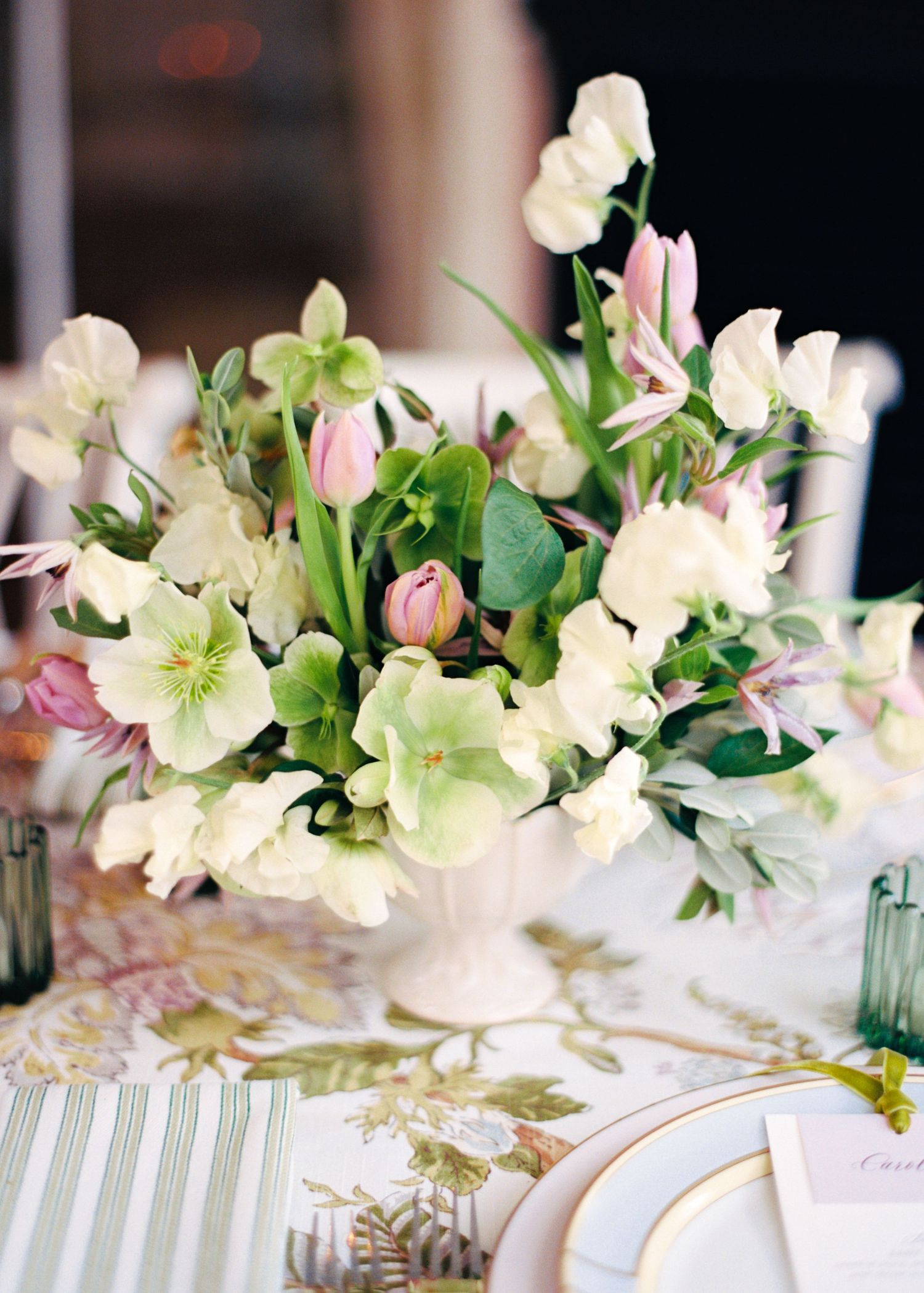 White pedestal vase with lush spring floral arrangement featuring pink tulips, hellebores, and sweet peas on an elegant tablescape.