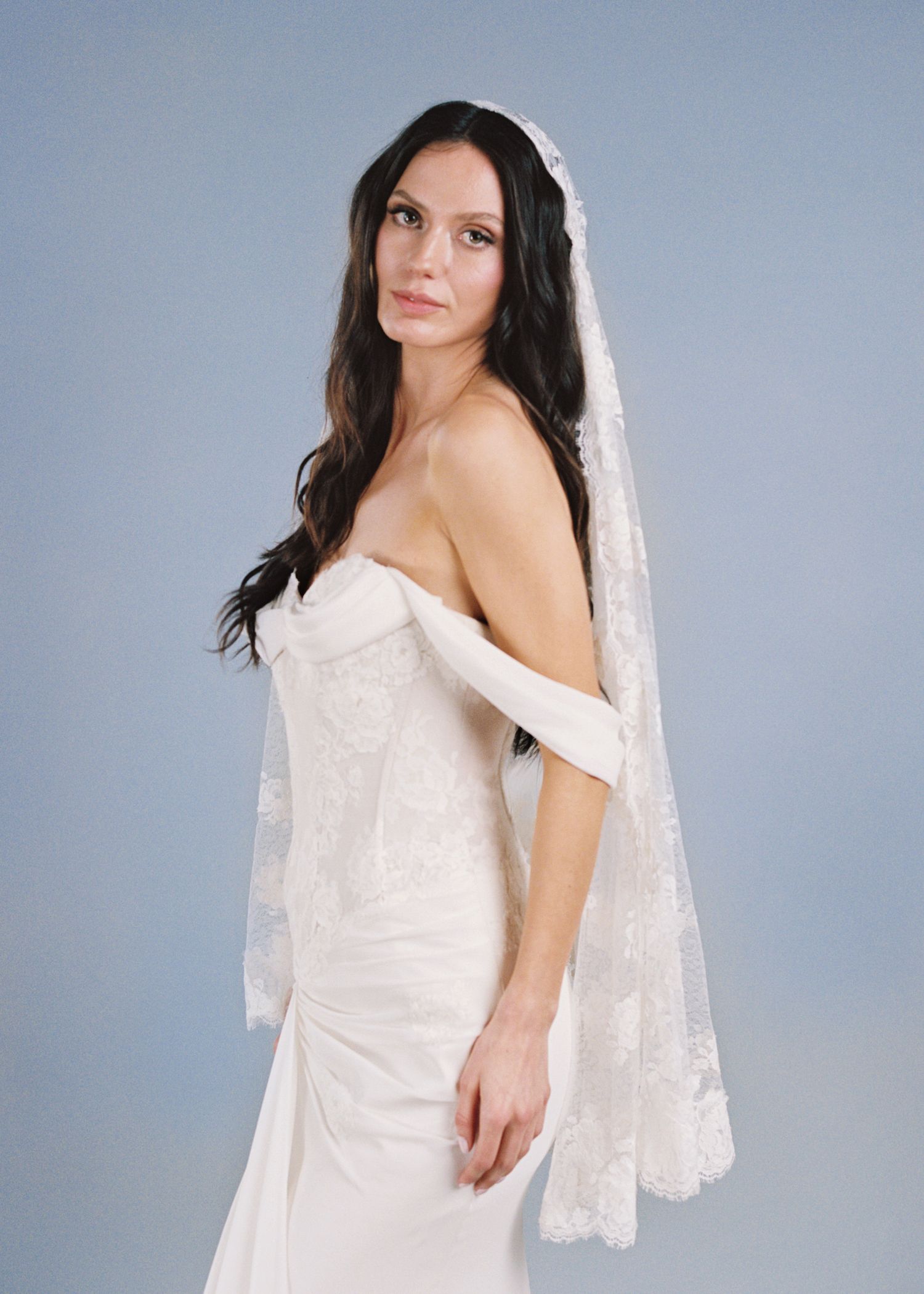 Bride in strapless white gown with lace mantilla veil, posing elegantly against a soft blue background.