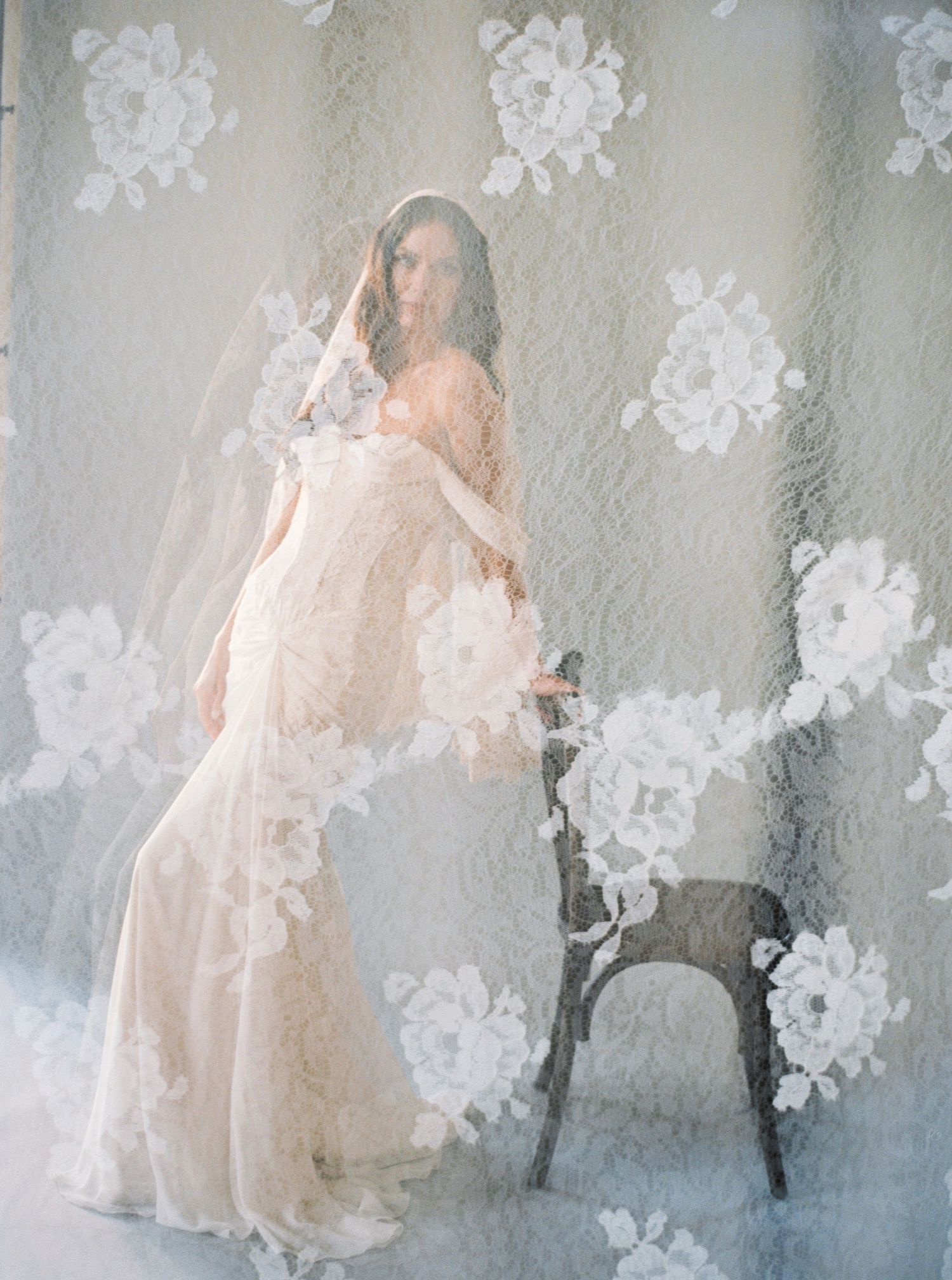 Bride in lace wedding gown poses gracefully beside a chair, draped in a floral veil against a soft floral backdrop.