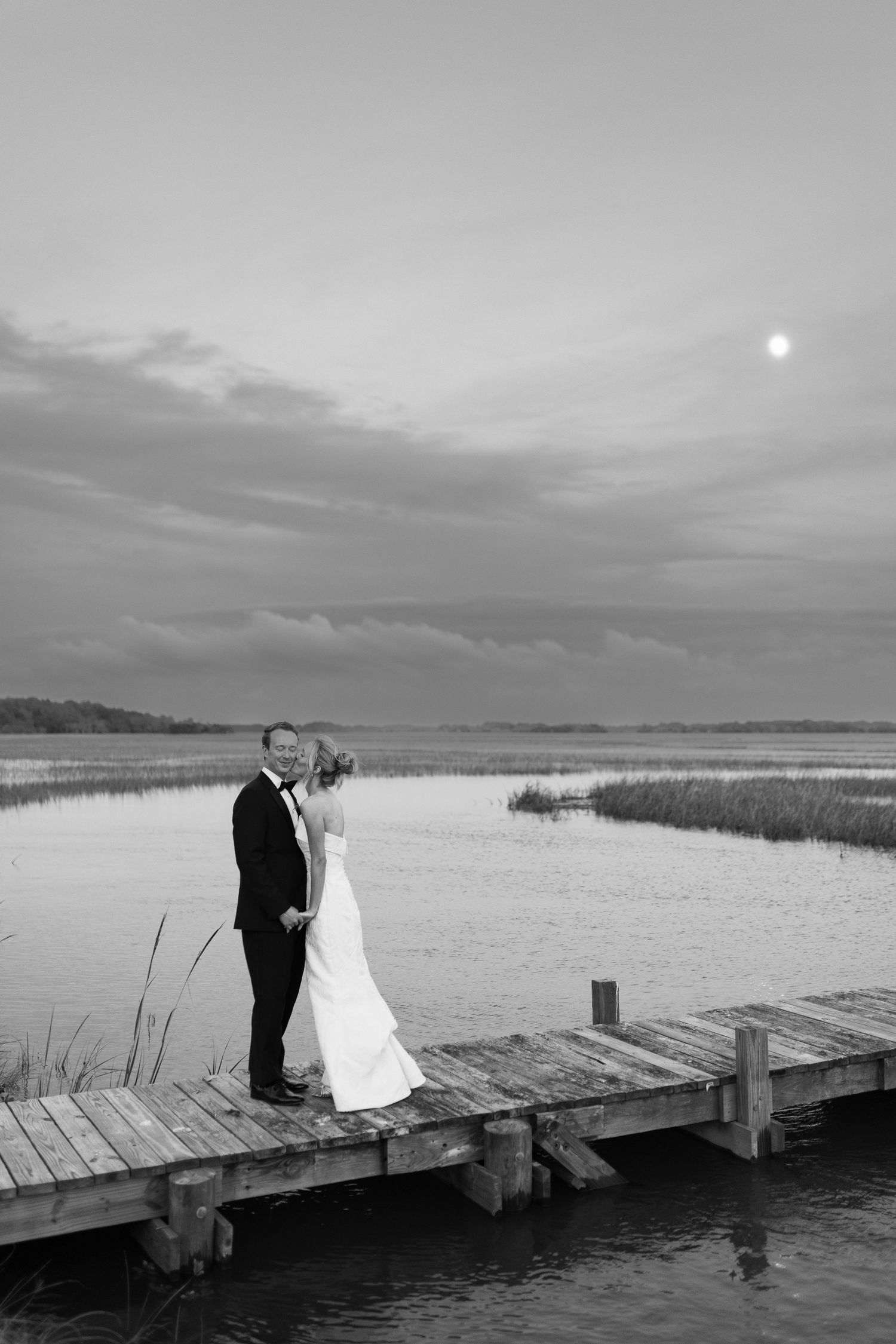 Wedding couple standing on wooden dock at sunset overlooking marsh waters.
