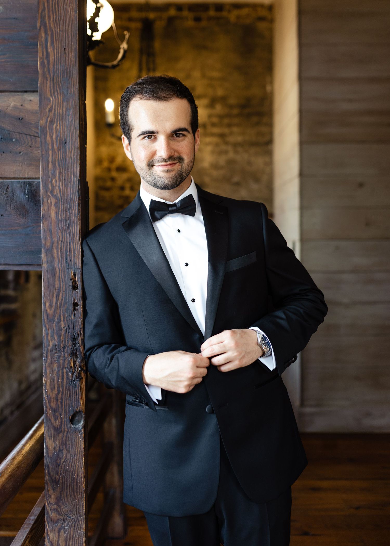 A groom in a black tuxedo with bow tie buttons his jacket while posing near a rustic wooden beam.