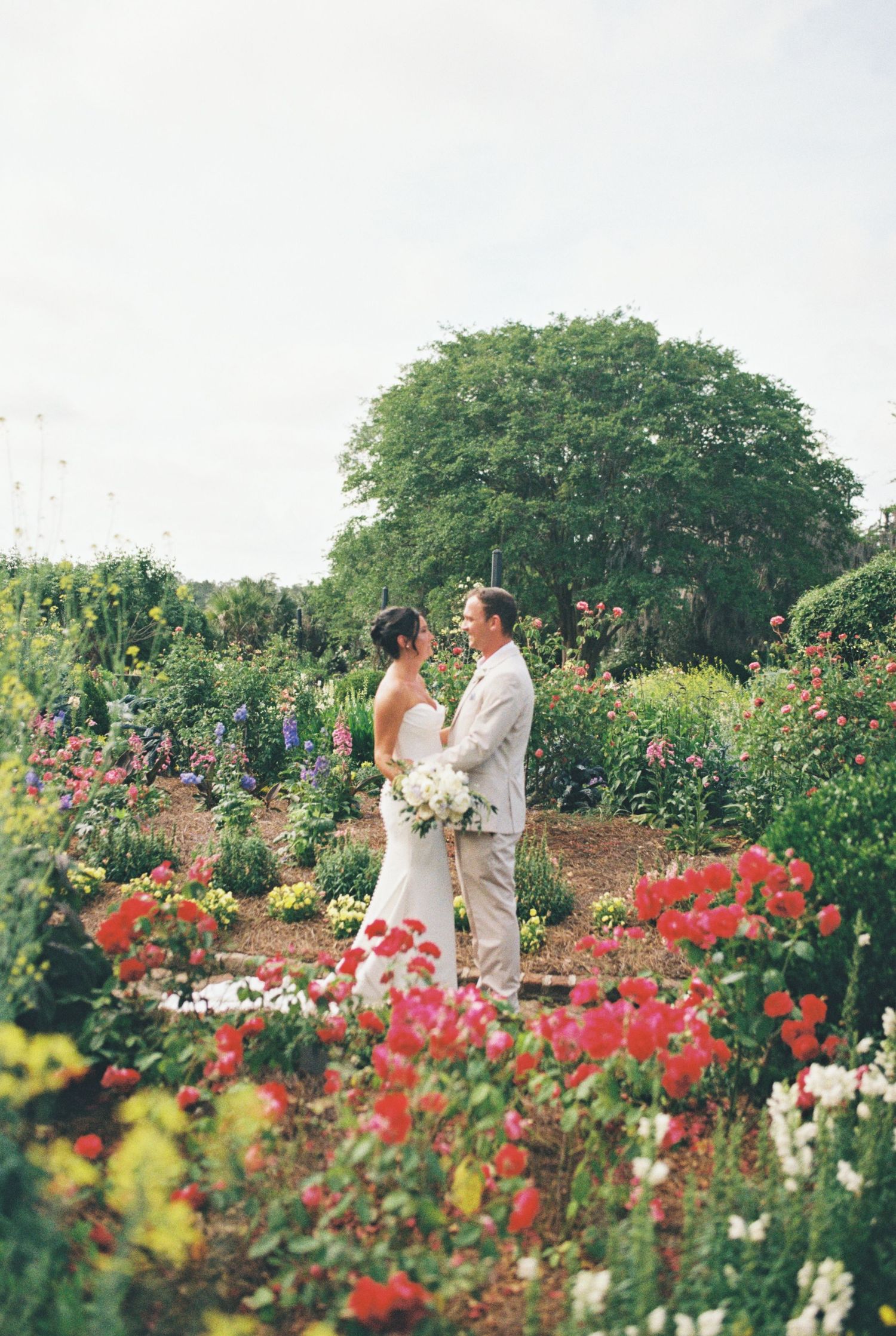 A bride and groom share a romantic moment surrounded by vibrant colorful flowers in a lush garden wedding setting.