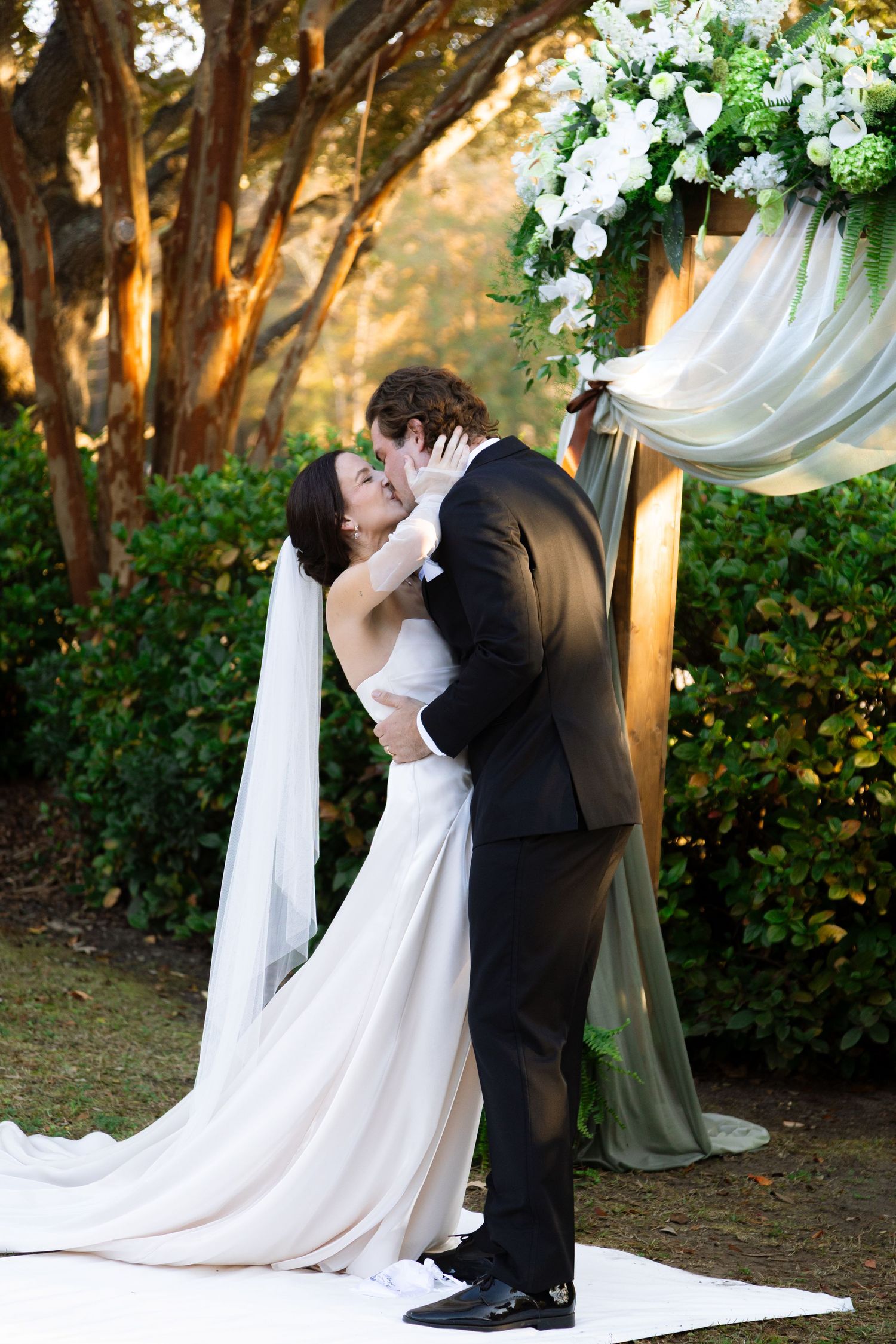 Romantic wedding ceremony under floral arch with white draping and trees in background.