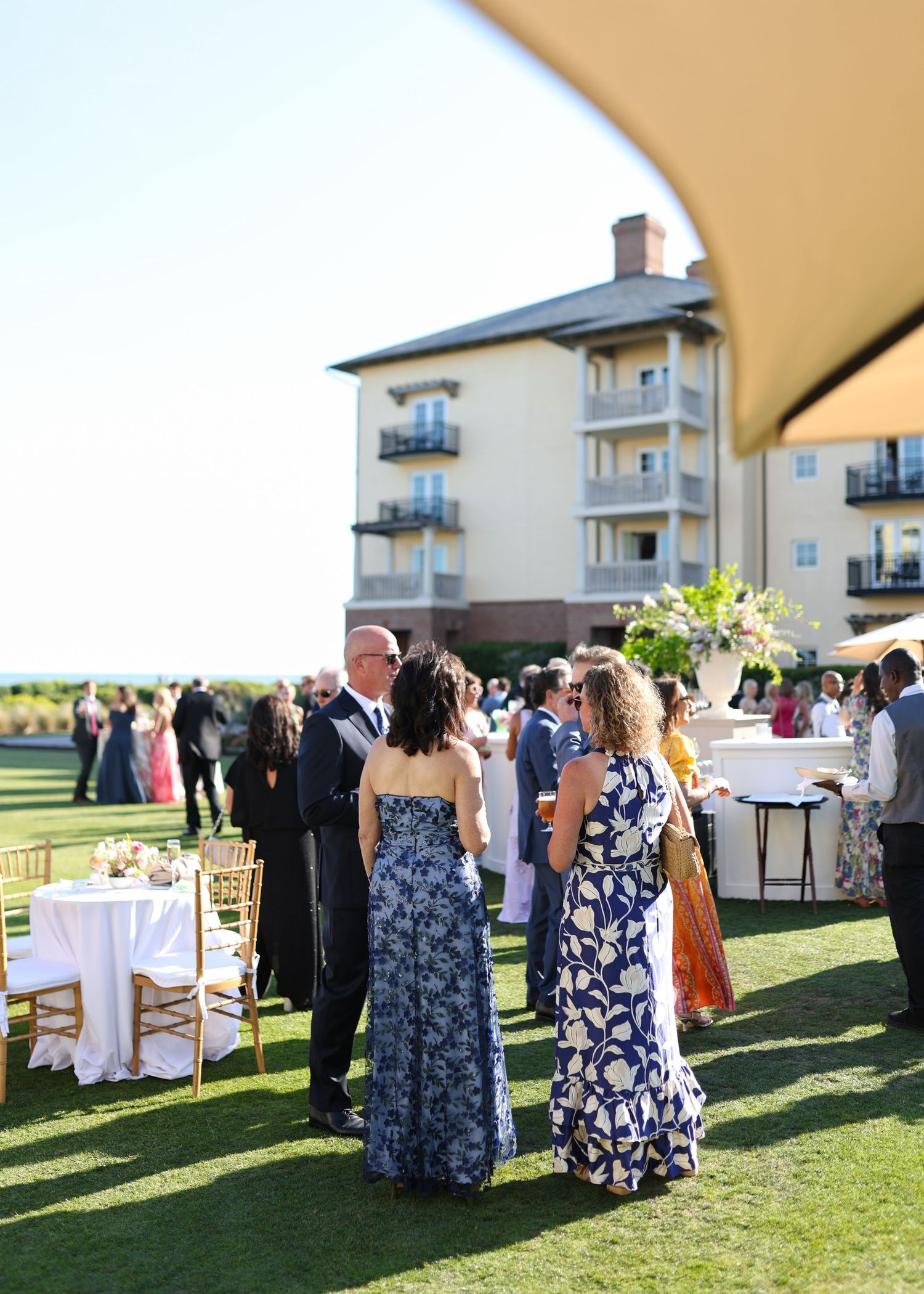 Elegant outdoor wedding reception on green lawn with guests in formal attire, gold chairs, and a multi-story venue building backdrop.