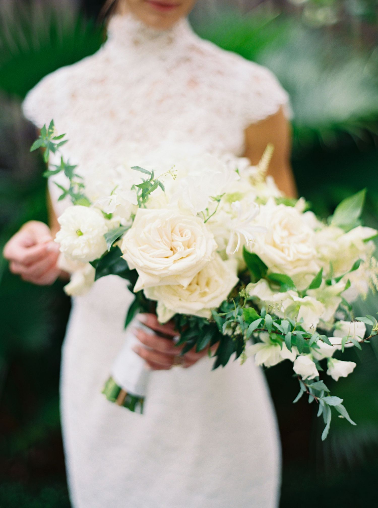 Bride in lace high-neck gown holding elegant white and cream rose bouquet with greenery at outdoor wedding.