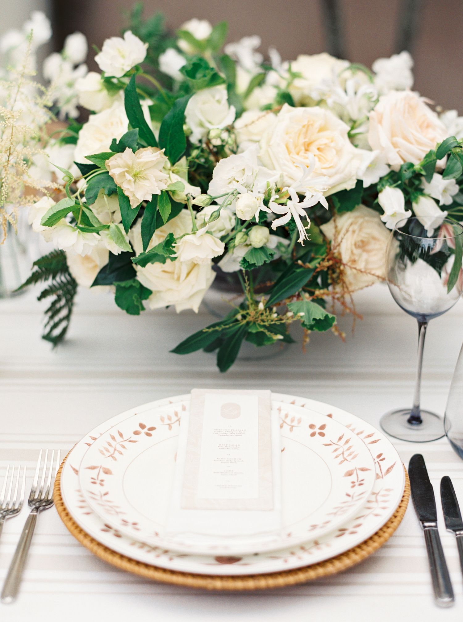 Elegant wedding table setting with white floral centerpiece, gold-rimmed charger plate, and menu card.