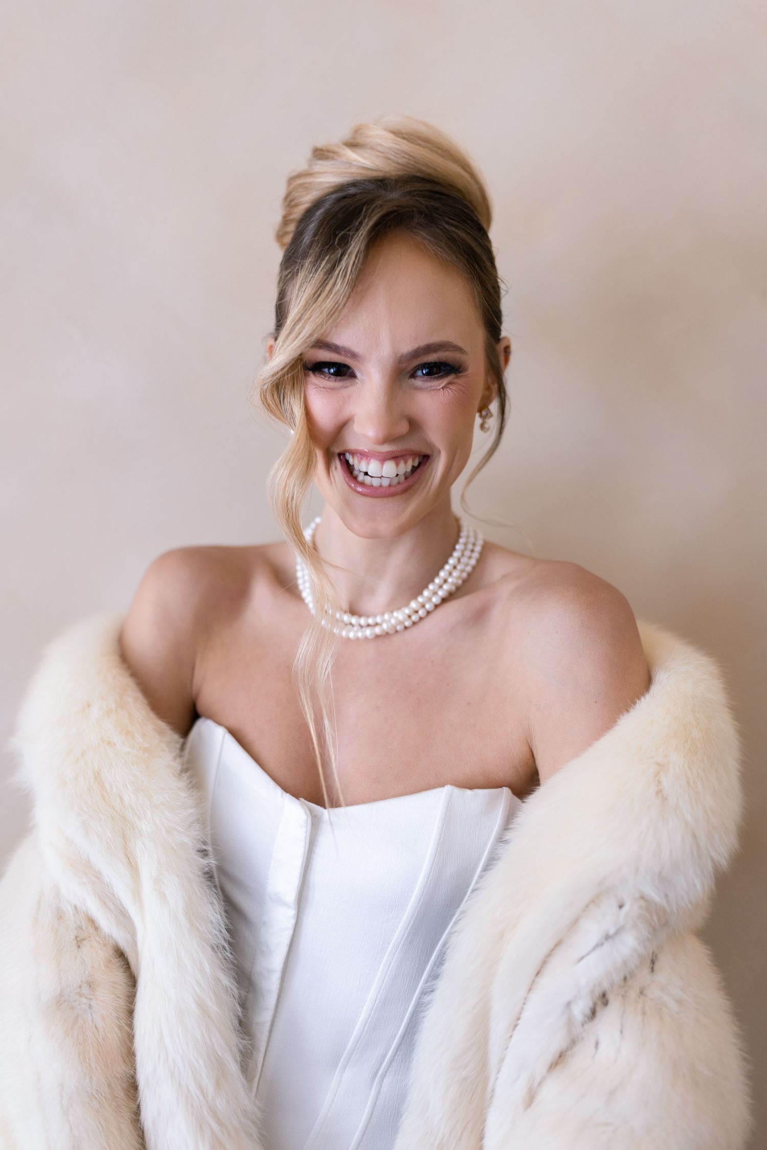 Smiling bride in white strapless gown, pearl necklace, and ivory fur stole with elegant updo hairstyle.