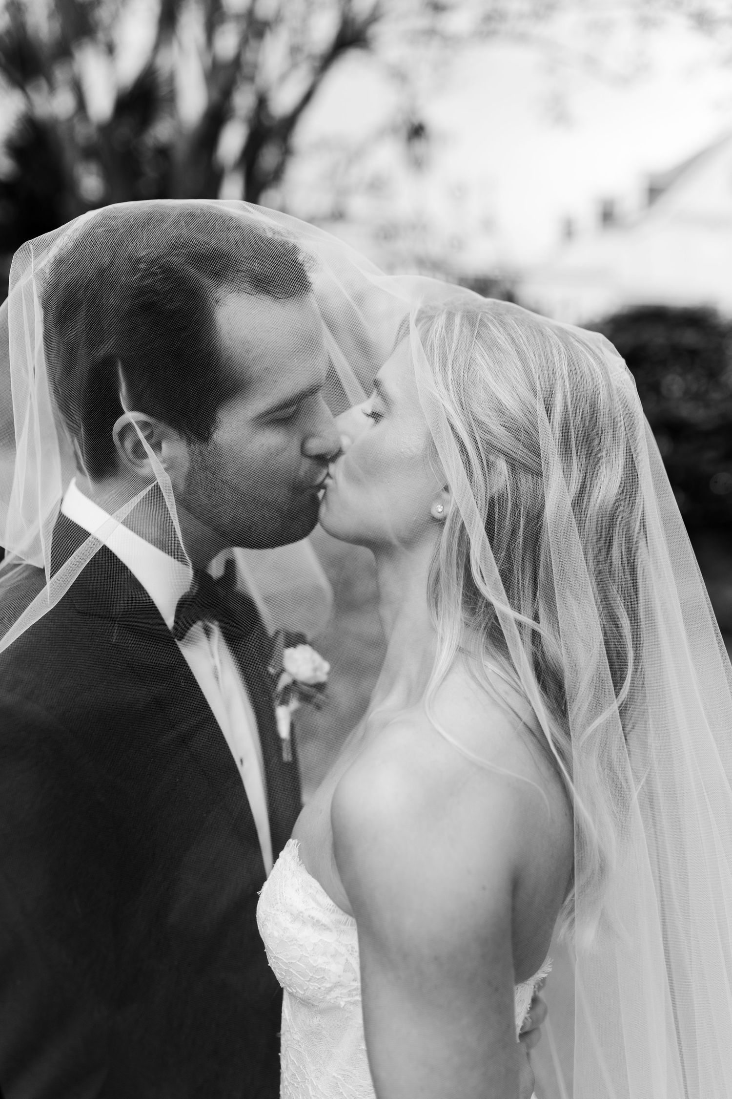 Black and white photo of a bride and groom sharing a romantic kiss under a flowing veil on their wedding day.