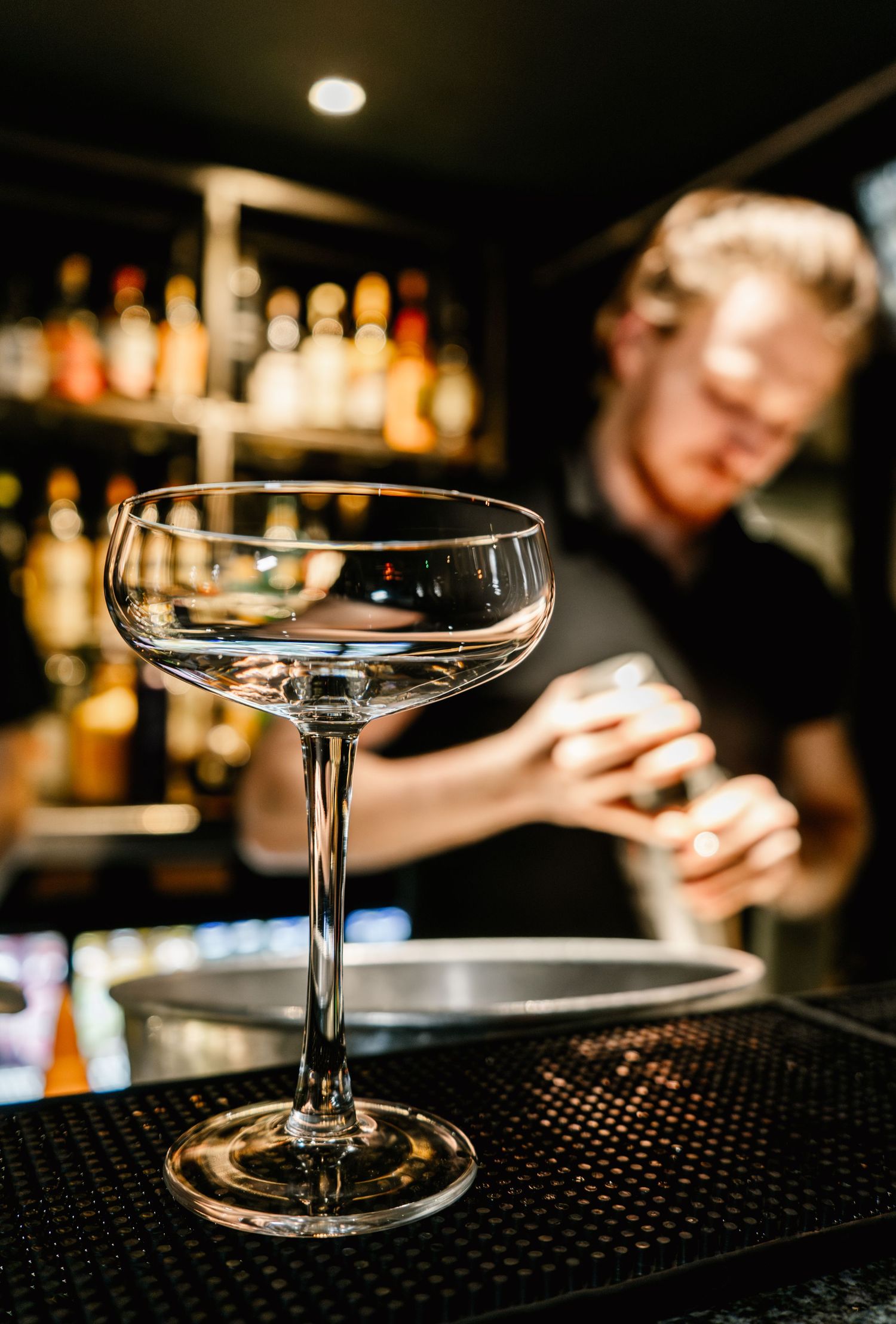 Elegant coupe glasses lined up on a dark bar counter with bottles in background.