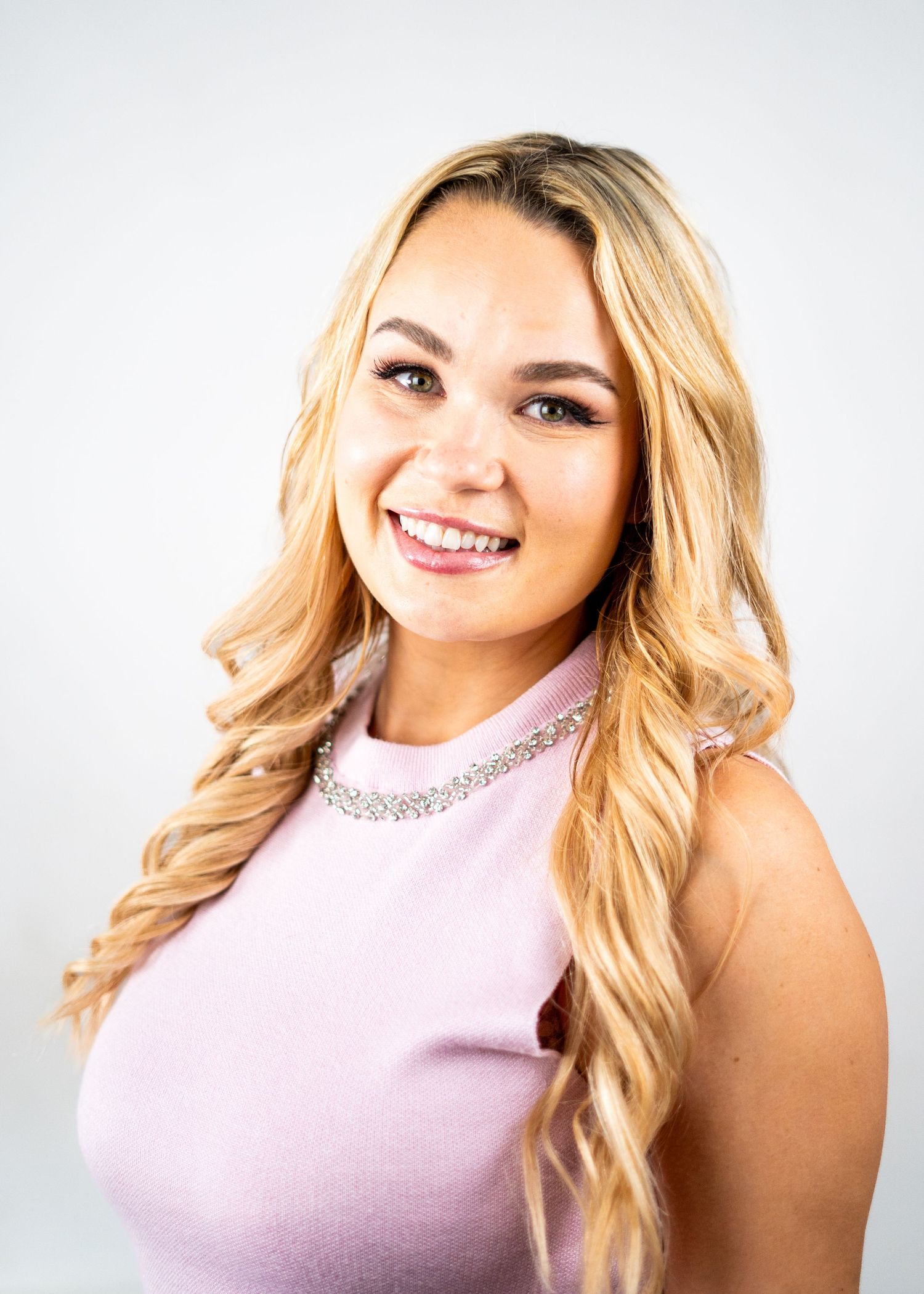 Professional portrait of a person in a pink top with long wavy blonde hair against a white backdrop.