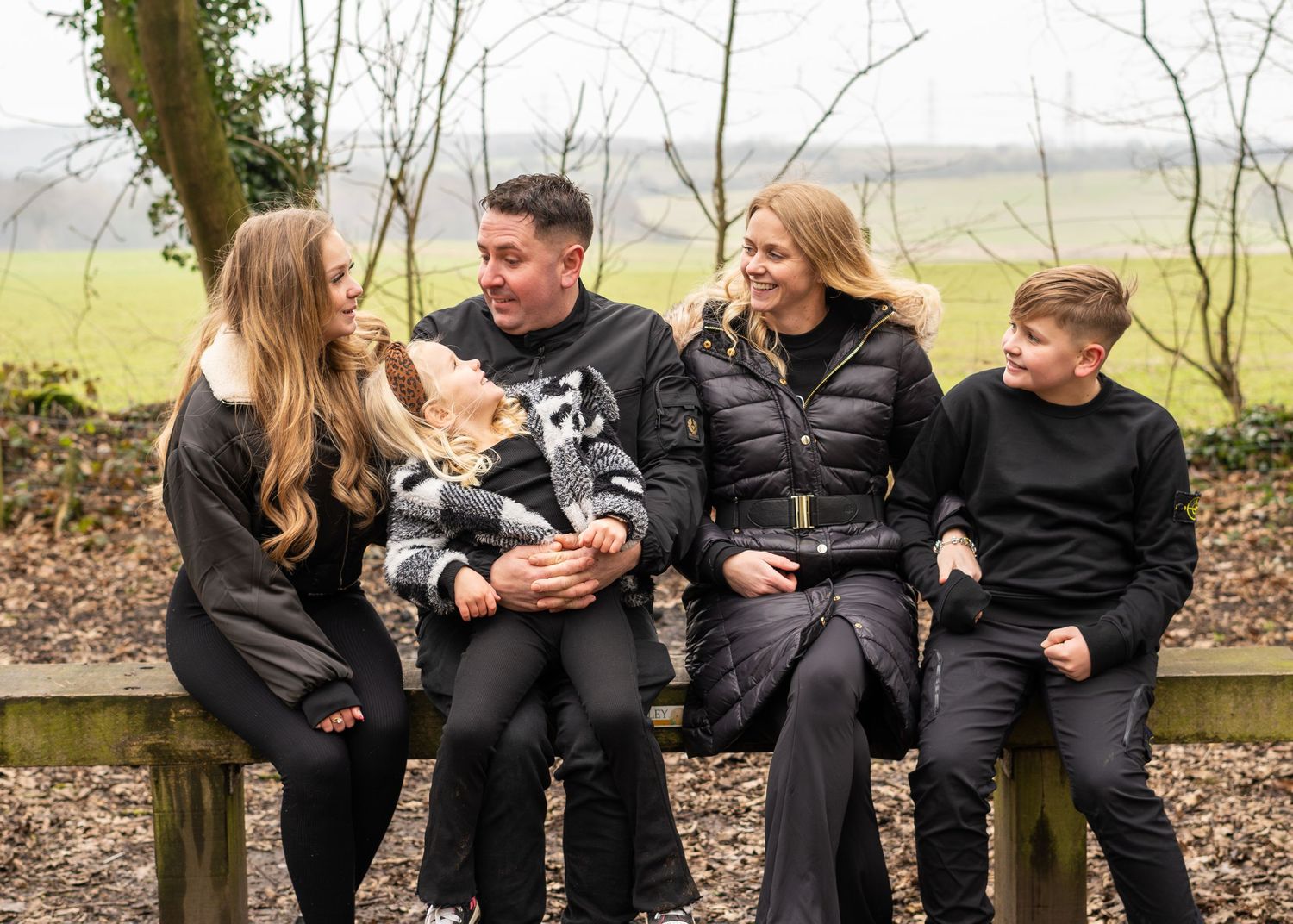 A family wearing black sits together on a wooden bench in a natural outdoor setting.