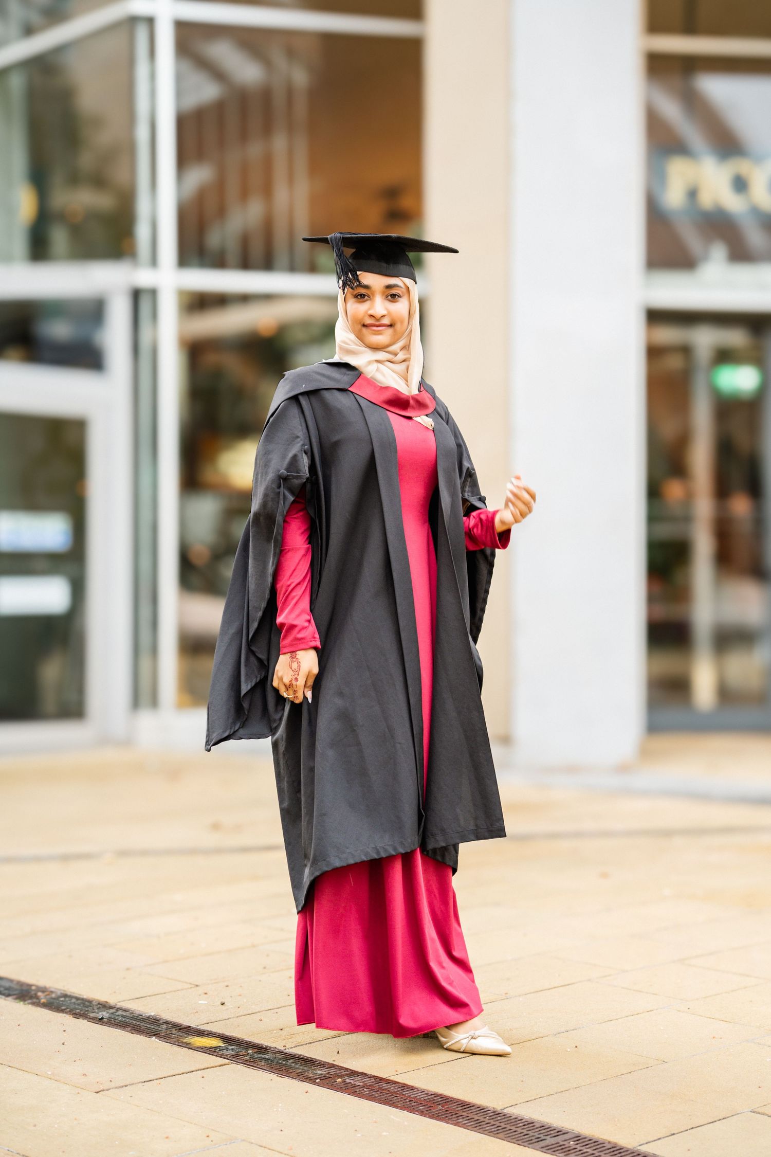 Graduate wearing black graduation gown and red dress poses near campus entrance.