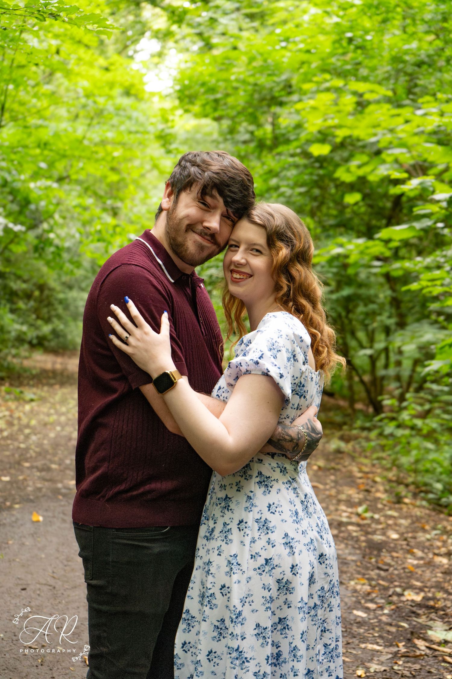 A romantic couple embraces on a forest path surrounded by lush green foliage.