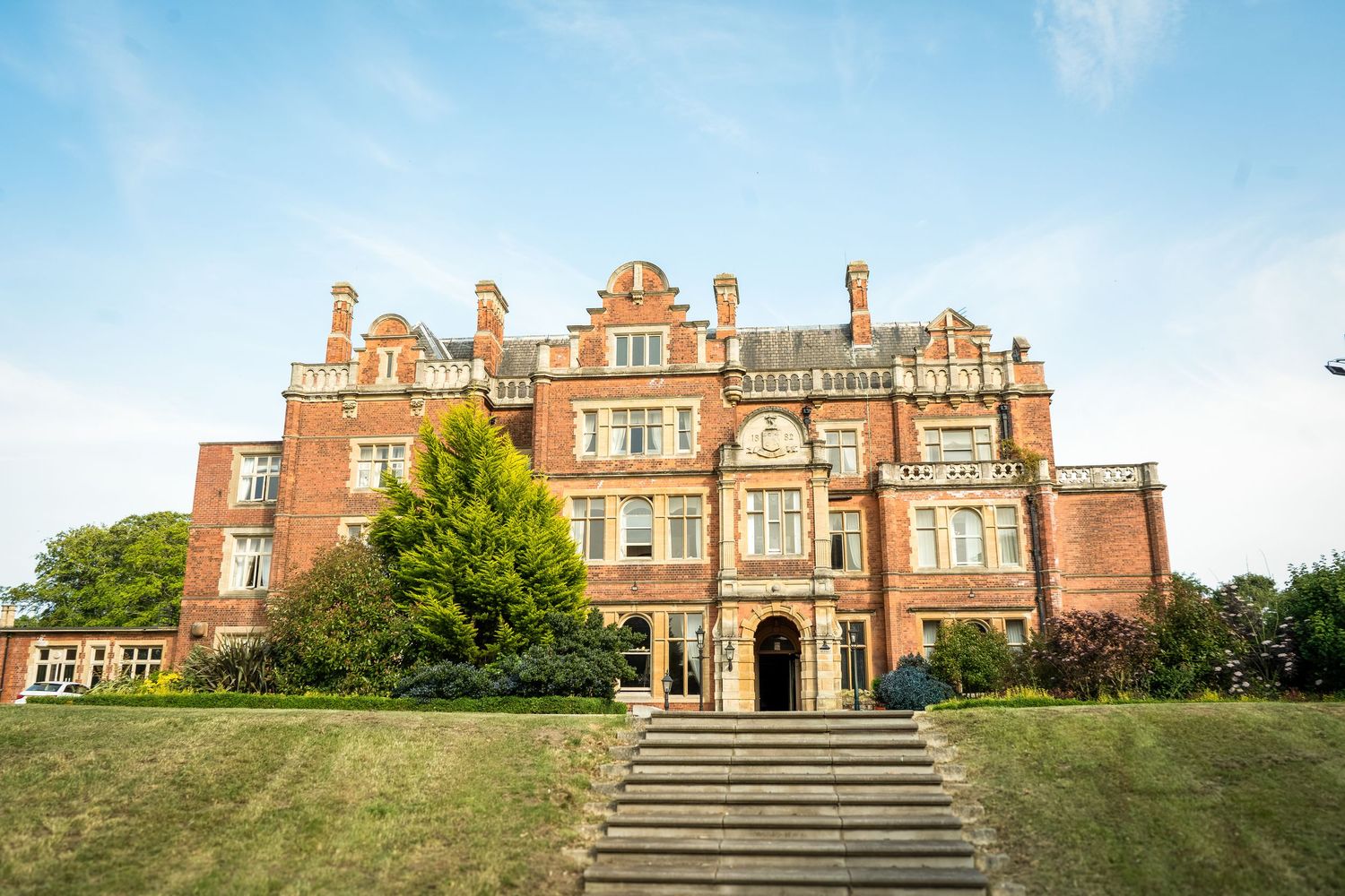 Panoramic view of a majestic red brick manor house with grand entrance steps and landscaped grounds.
