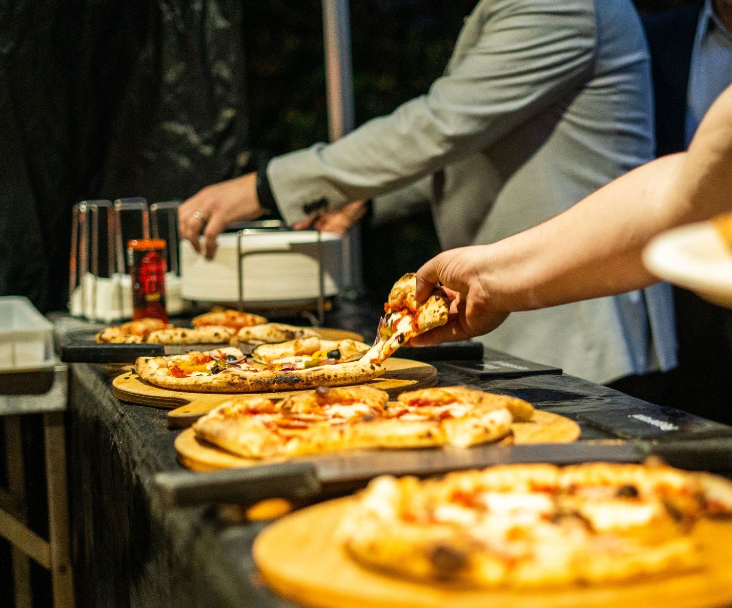 Fresh pizzas being prepared on wooden boards at an outdoor cooking station.