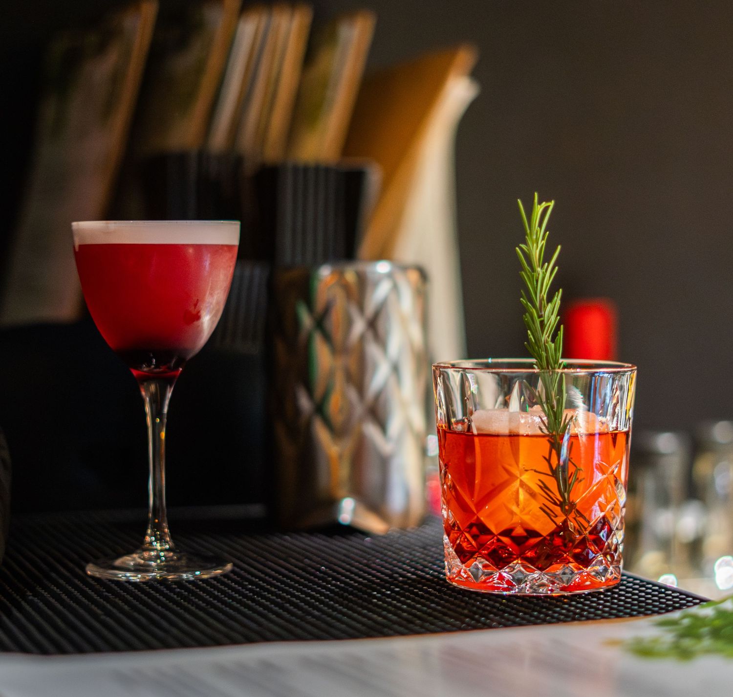 Red craft cocktails garnished with rosemary on a dark bar counter.
