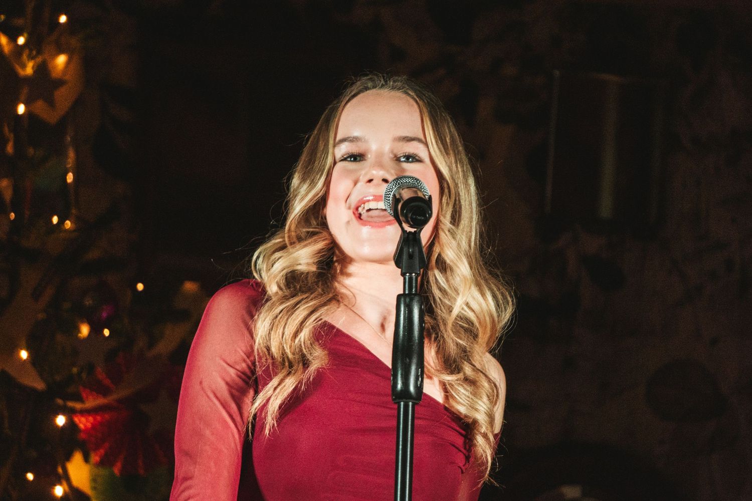 Performer in burgundy dress sings into microphone on dimly lit stage.
