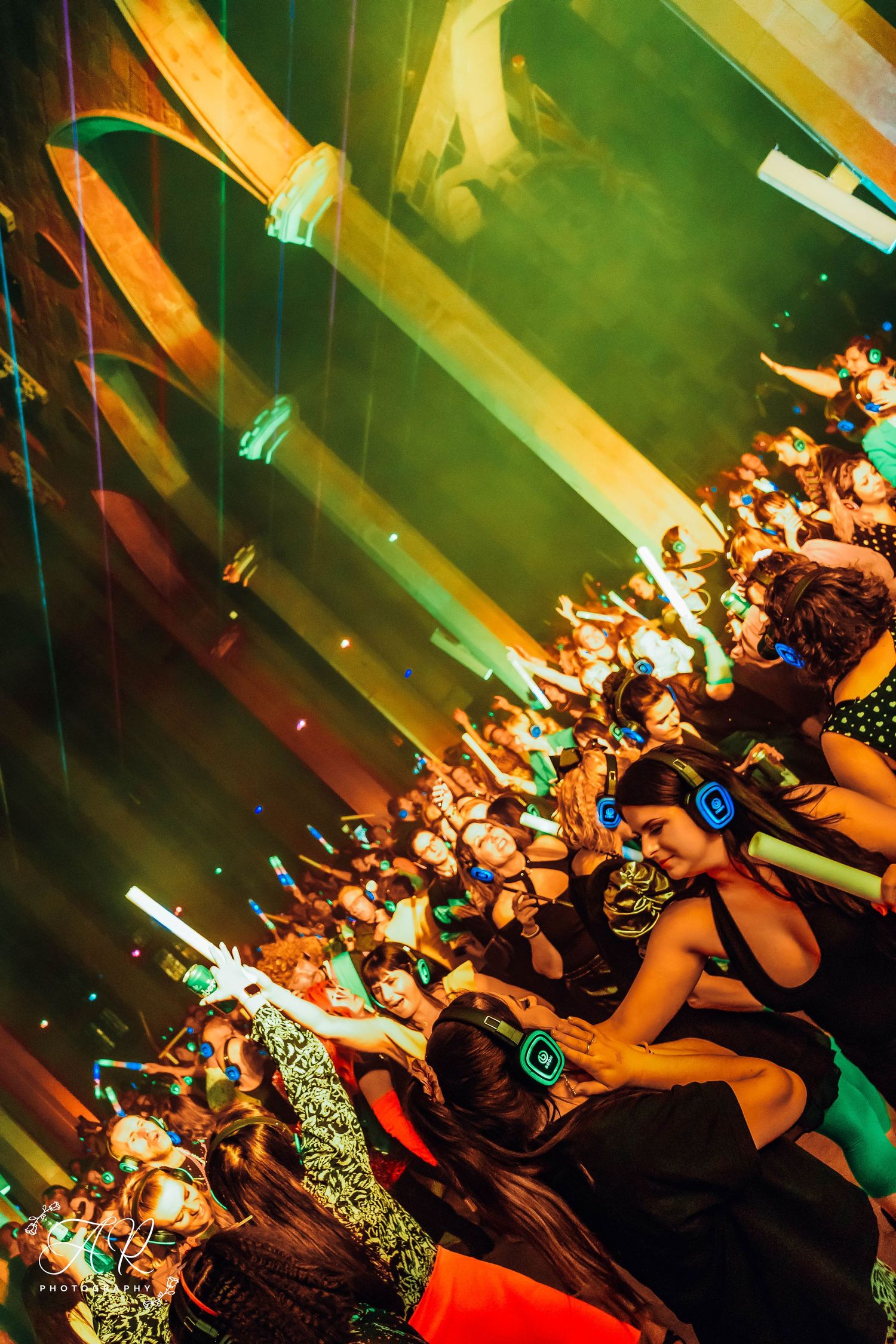 Dance floor filled with revelers under green and orange atmospheric lighting.