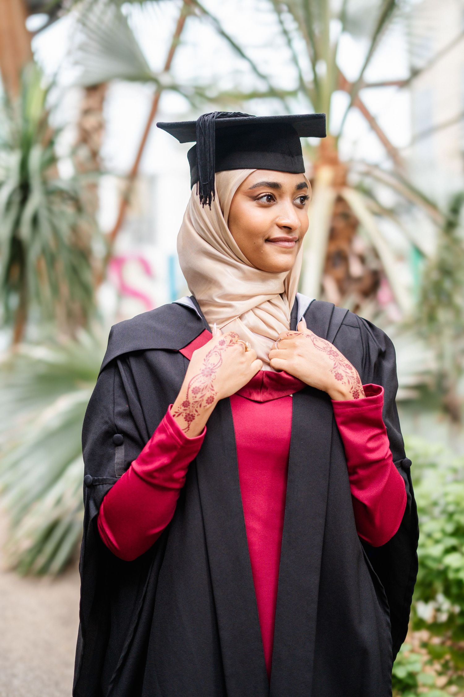 Graduate poses playfully in academic regalia against tropical greenhouse backdrop.