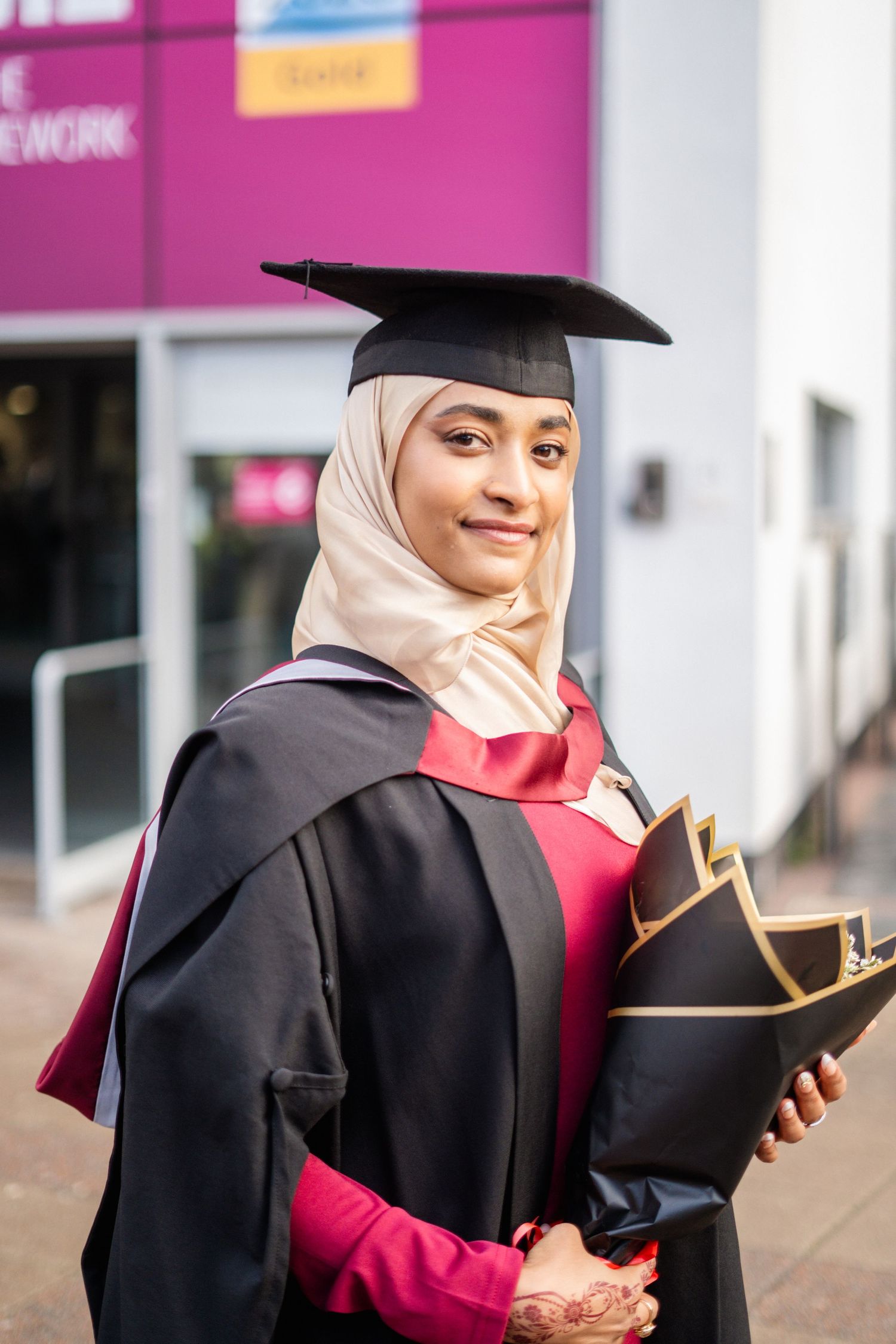 Graduate wearing hijab and academic regalia smiles while holding graduation materials.