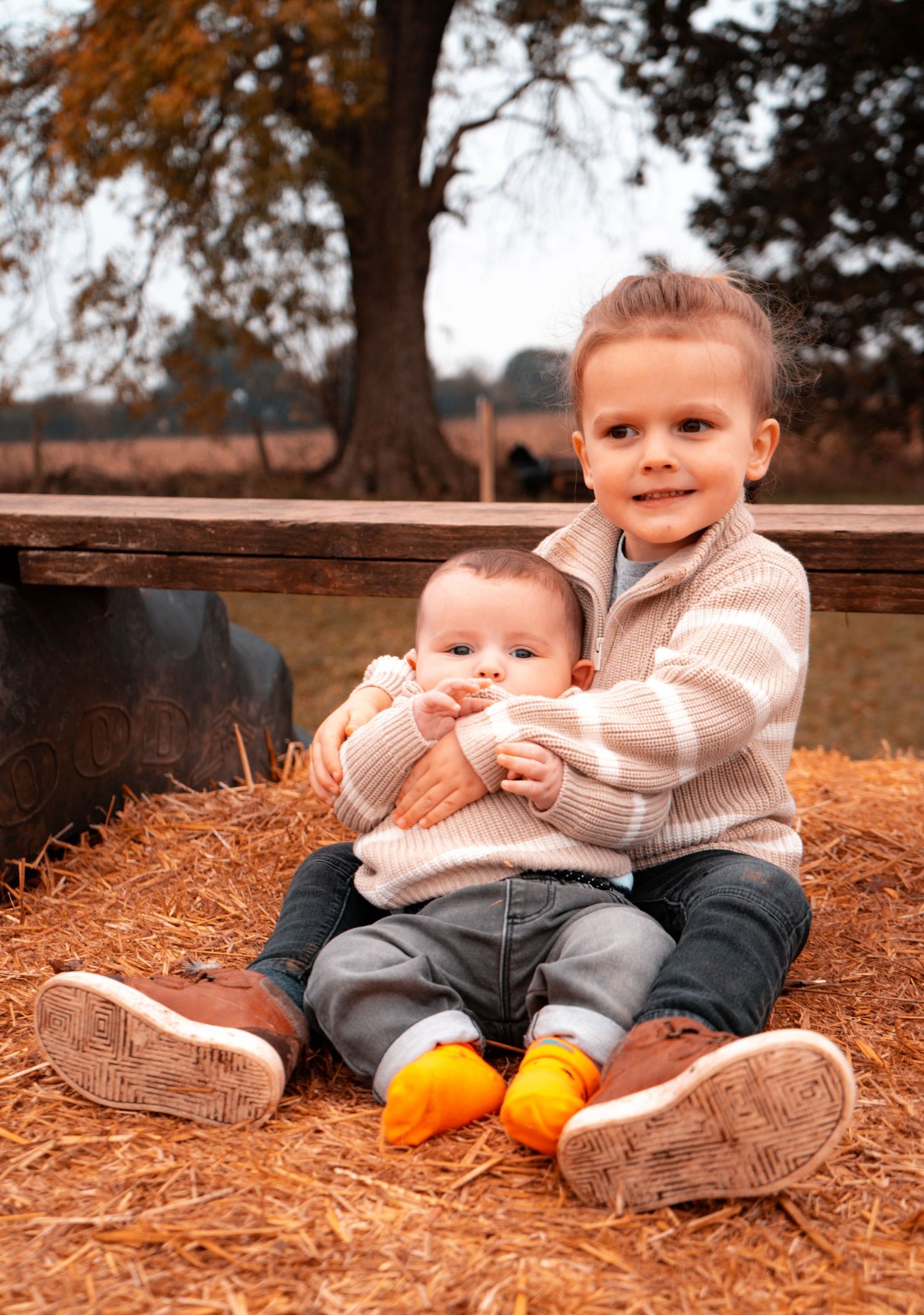A child sits on autumn leaves wearing a beige sweater and orange shoes.
