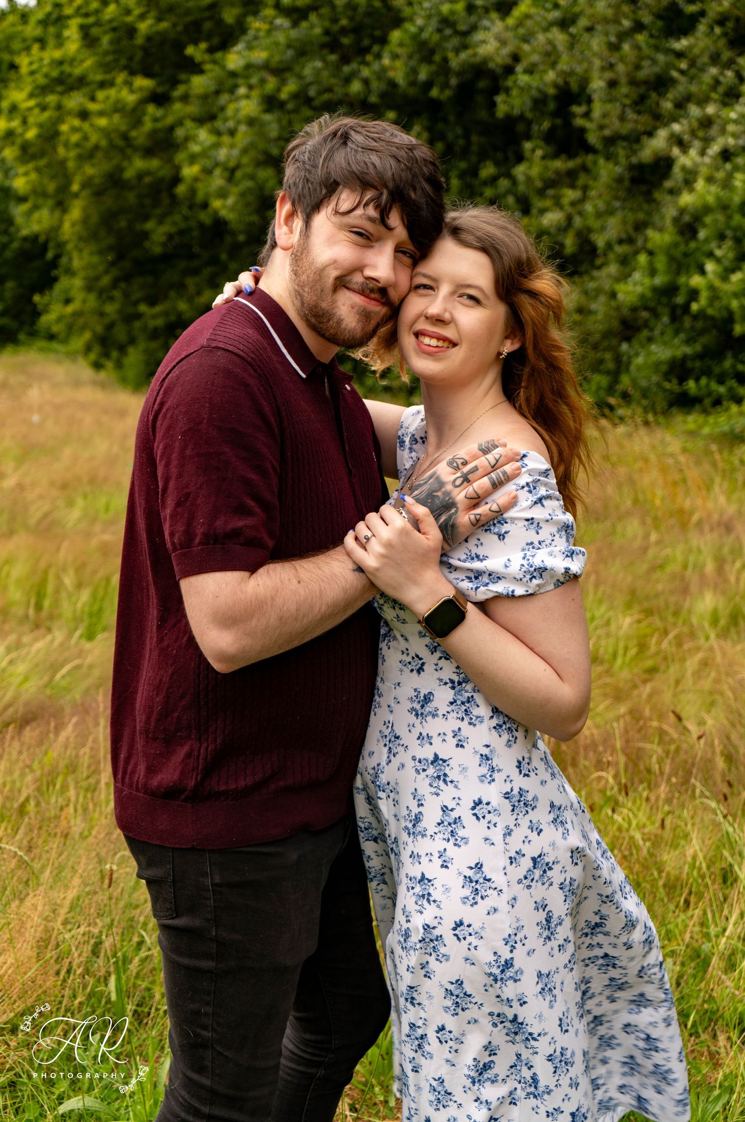 A couple shares joyful moments together in a sunny meadow with tall grass and trees.