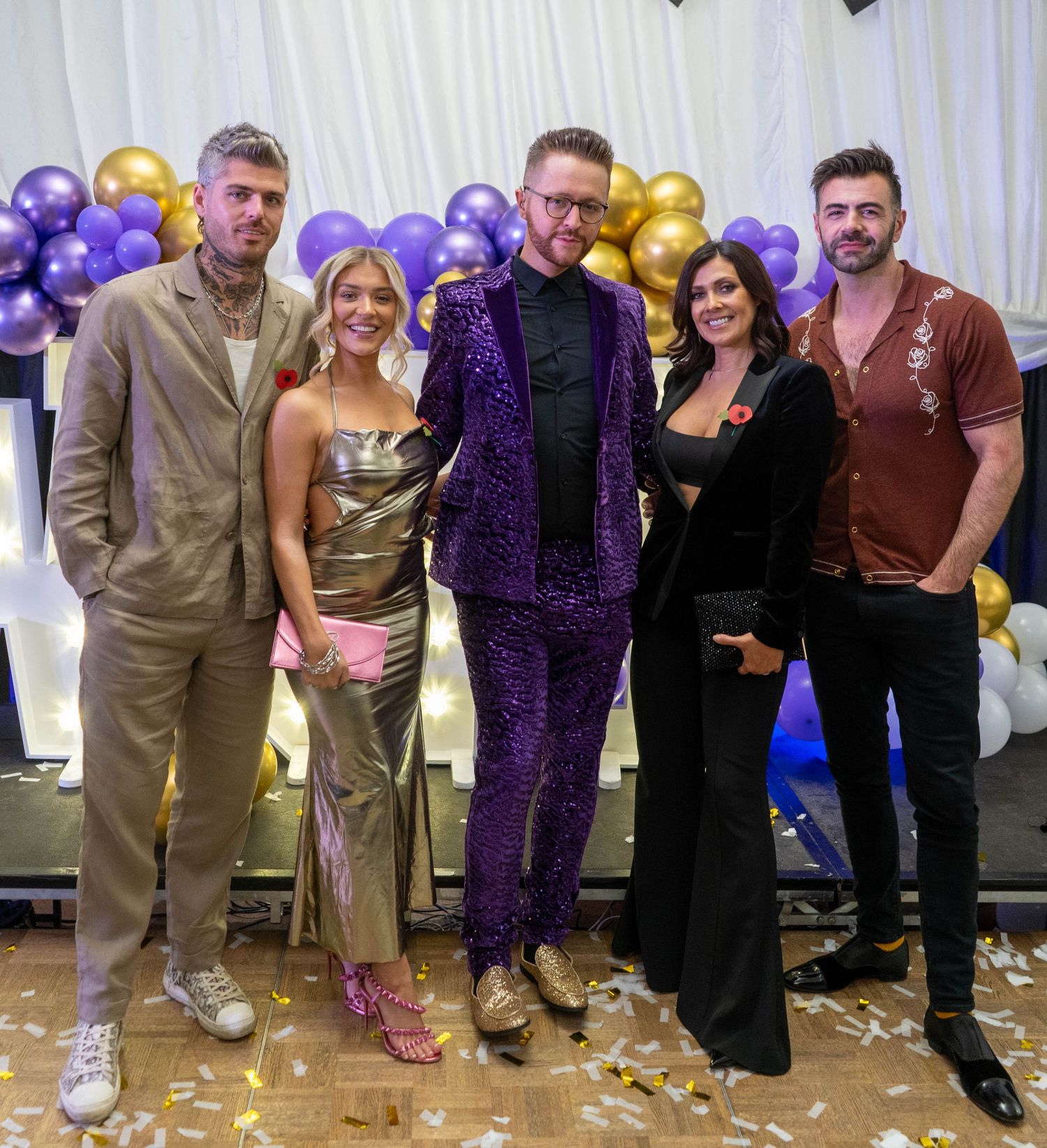 Group celebration photo with purple and gold balloon decorations and confetti on the floor.