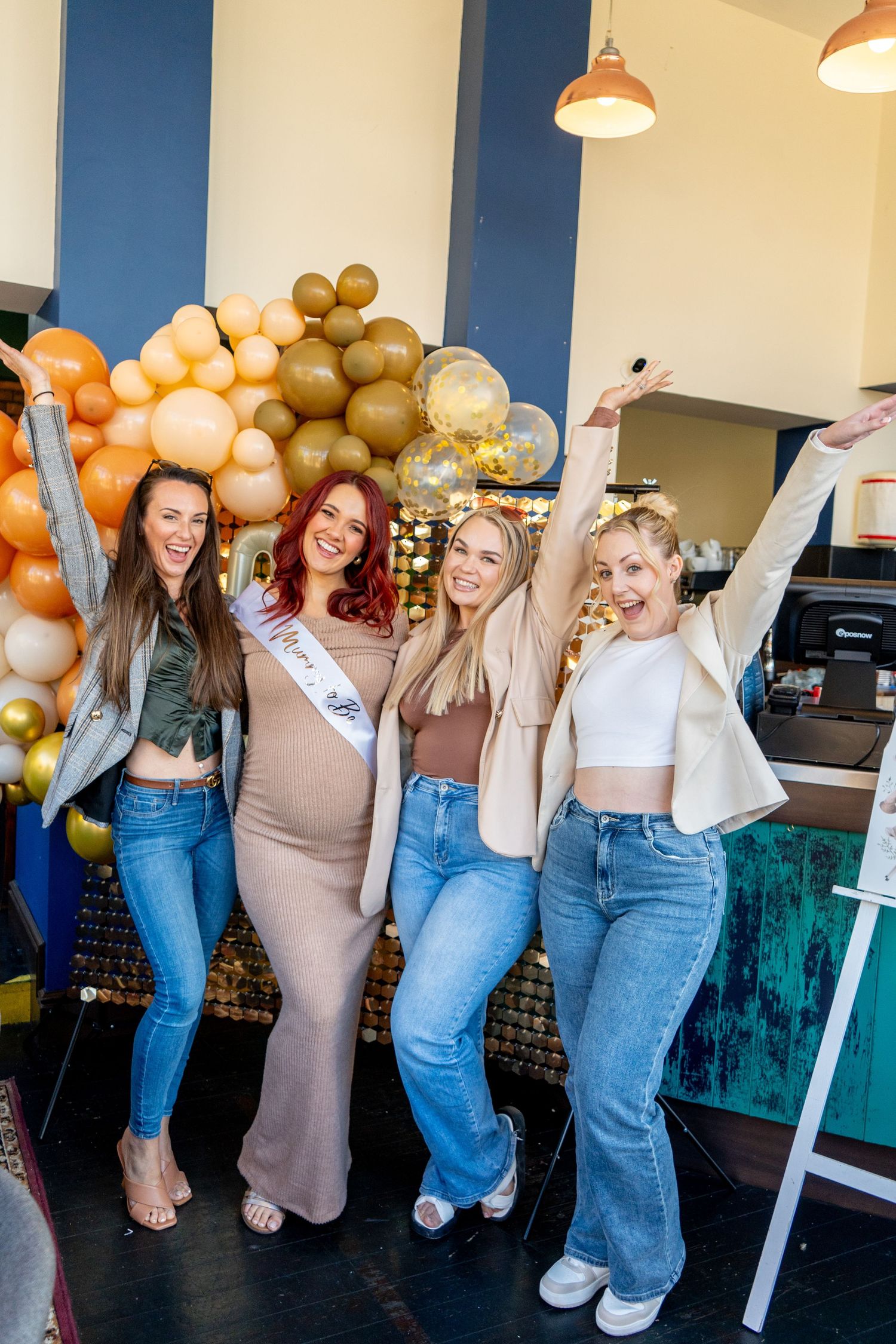 Two women in casual outfits pose playfully against a wall decorated with gold and orange balloons at a party.