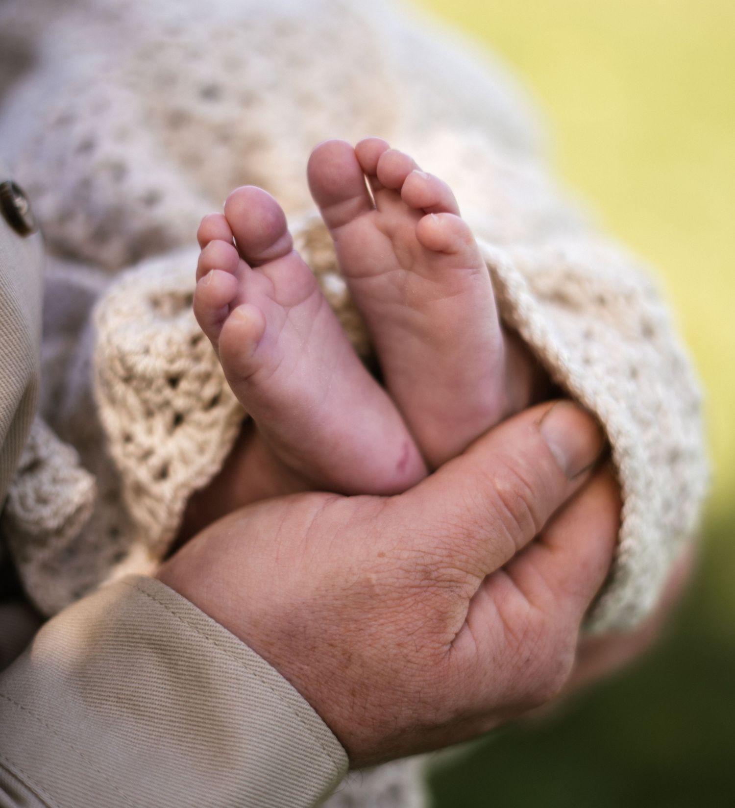 Tiny newborn feet peek out from underneath a cream crocheted blanket in sunlight.
