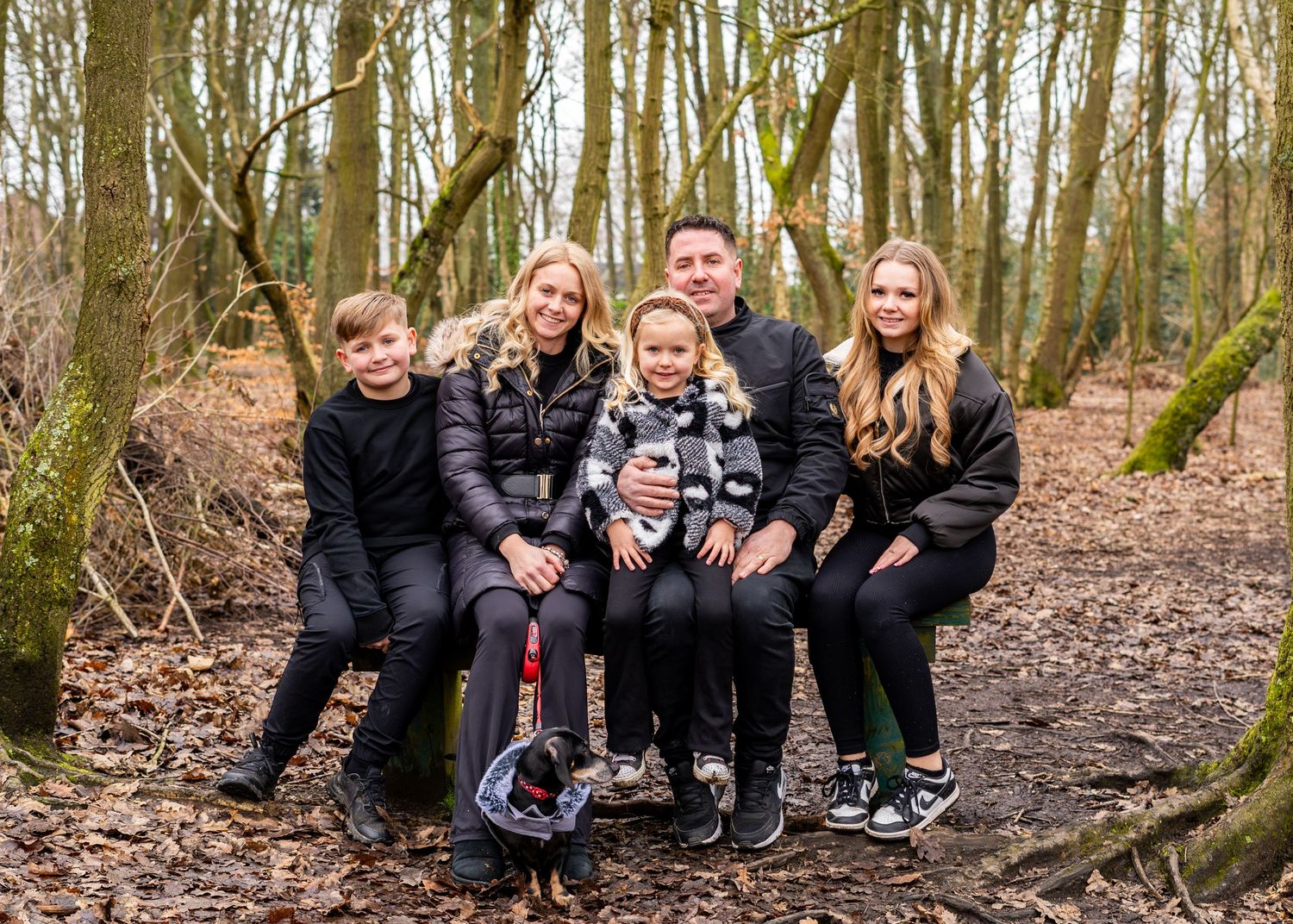 Family group portrait in wooded setting with fallen leaves and bare trees.