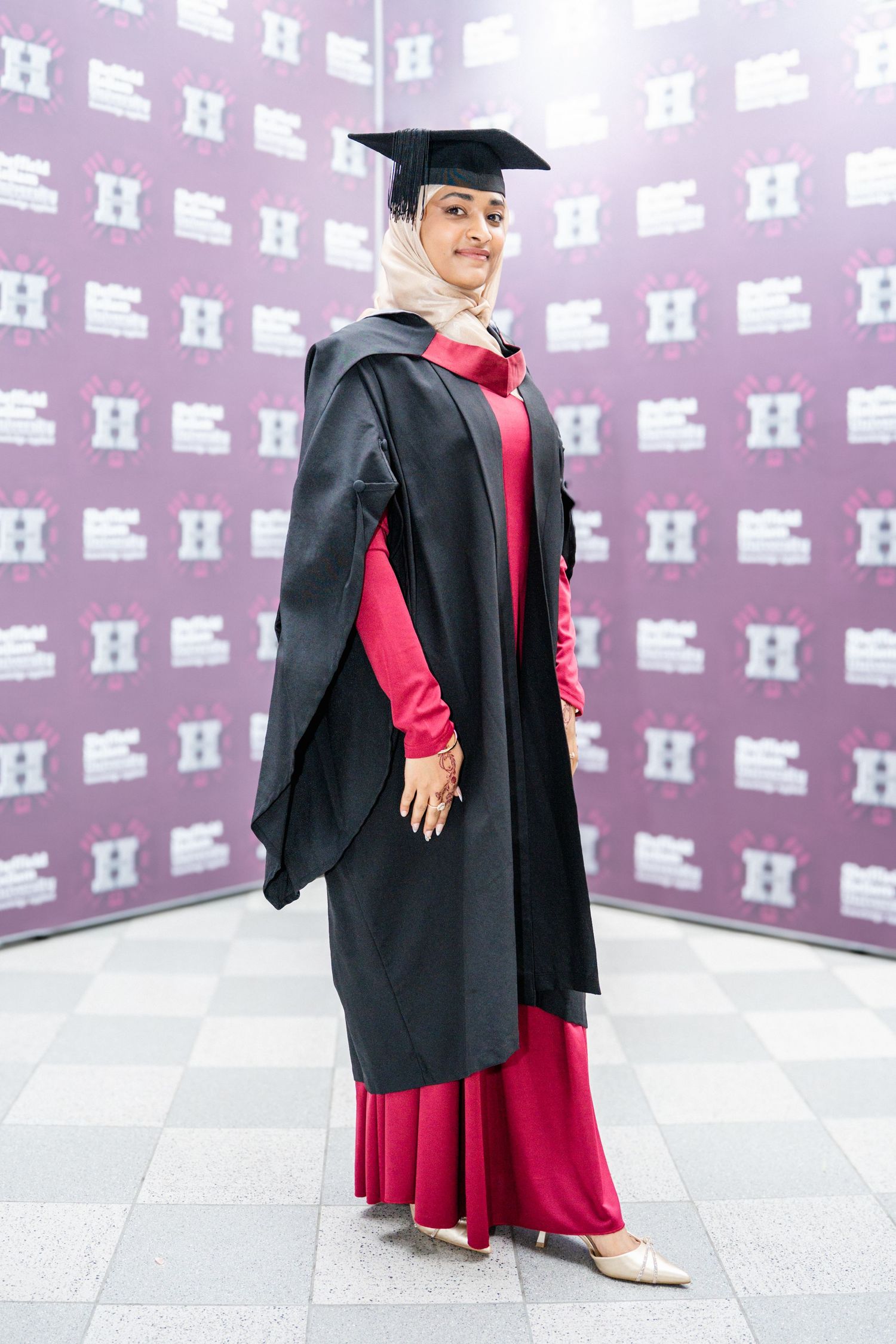 Graduate in black cap and gown with pink dress poses against patterned background during commencement ceremony.