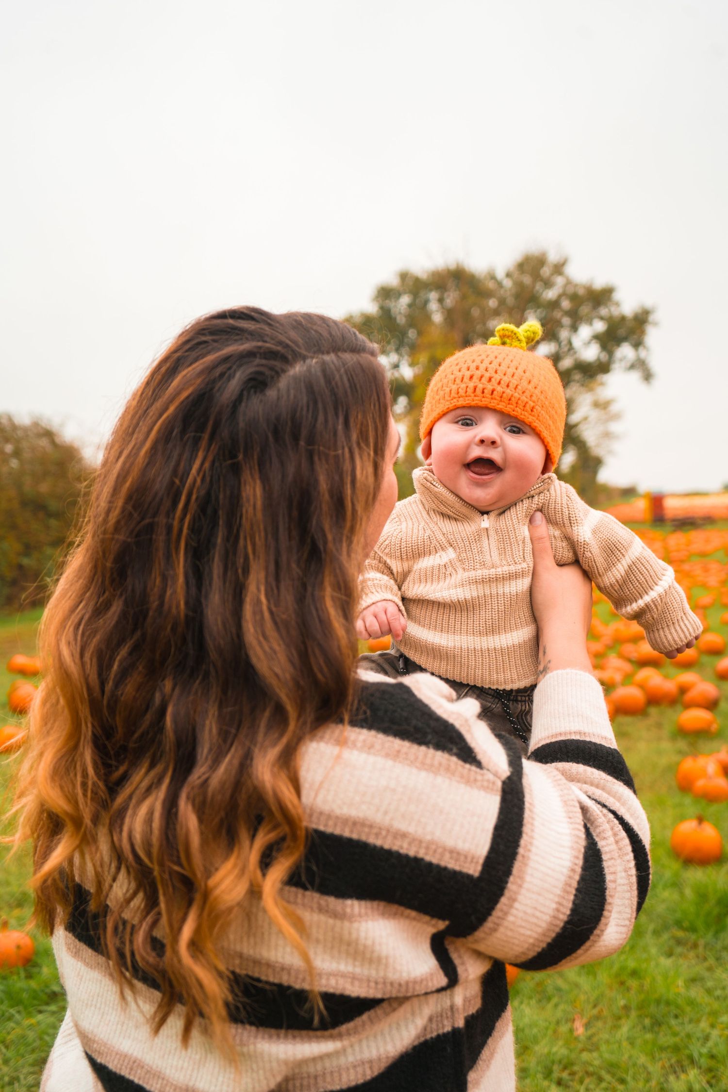 Parent holds smiling baby in orange hat at pumpkin patch during autumn.
