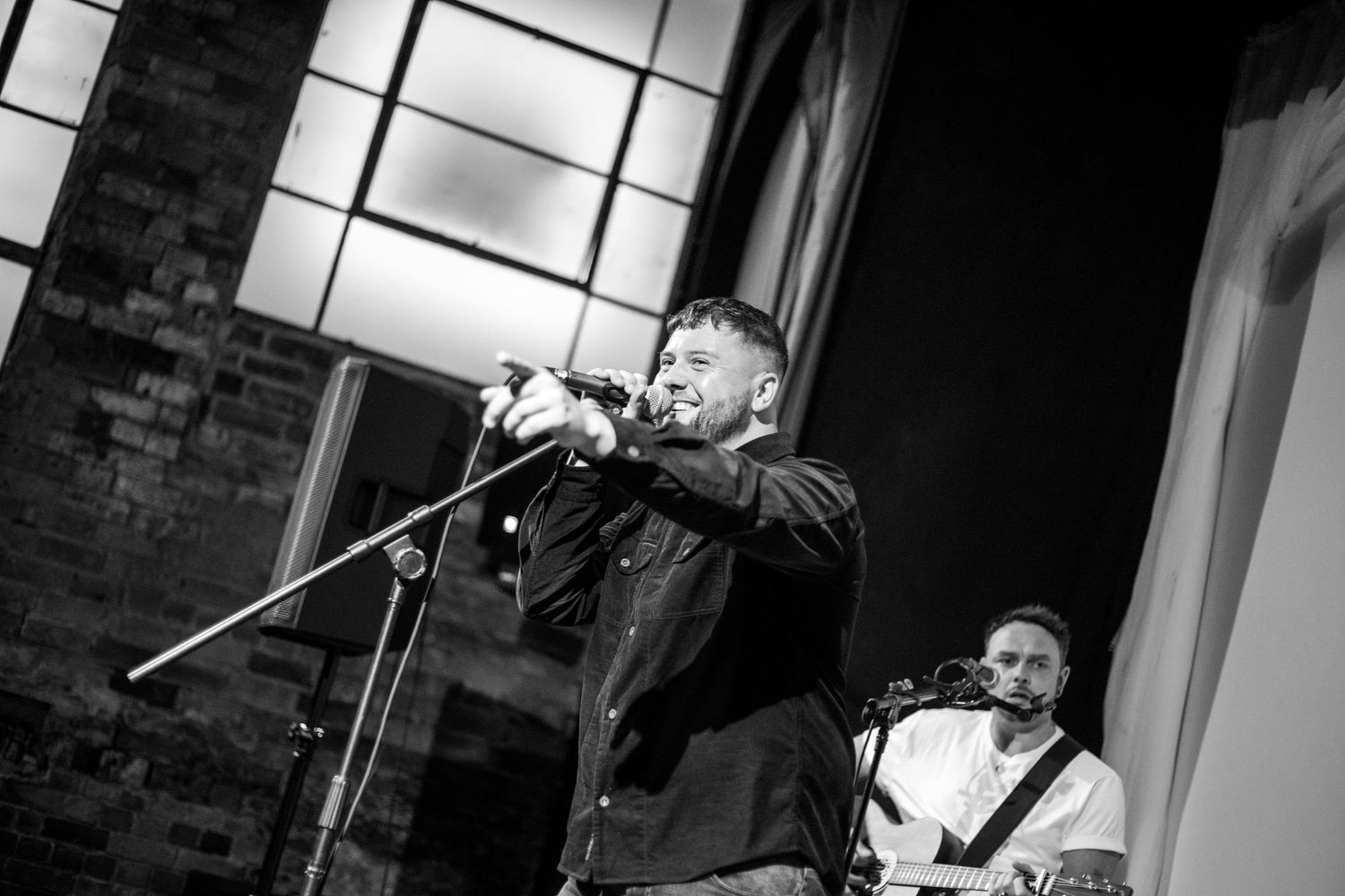 Black and white concert photo showing performers on stage with dramatic lighting and microphones.